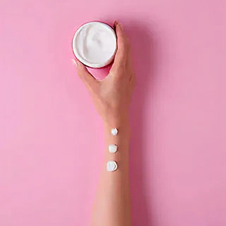 Hand holding a container of white cream with three dollops of cream on arm against pink background.