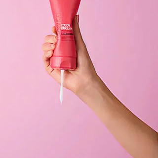 Hand holding a pink tube of color protectant hair product against a pink background.