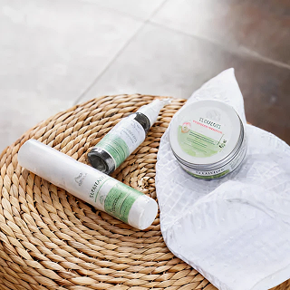 Skincare products, including a tube, a bottle, and a jar, placed on a woven mat with a white cloth underneath