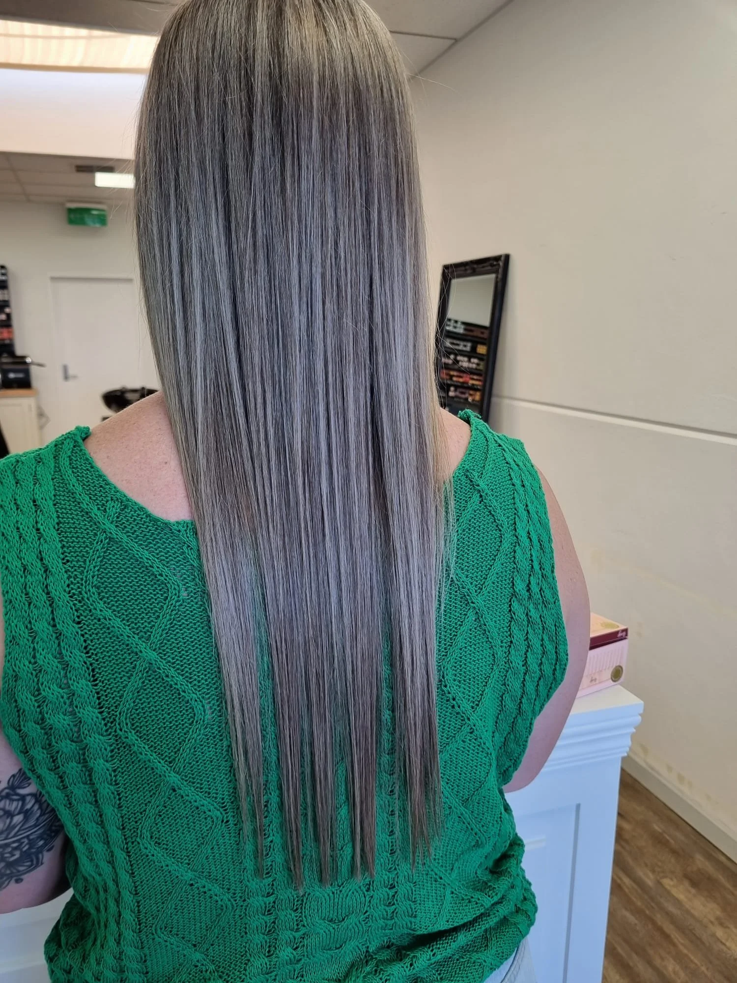 Person with long, straight, silver-gray hair wearing a green knit top, standing in a salon.