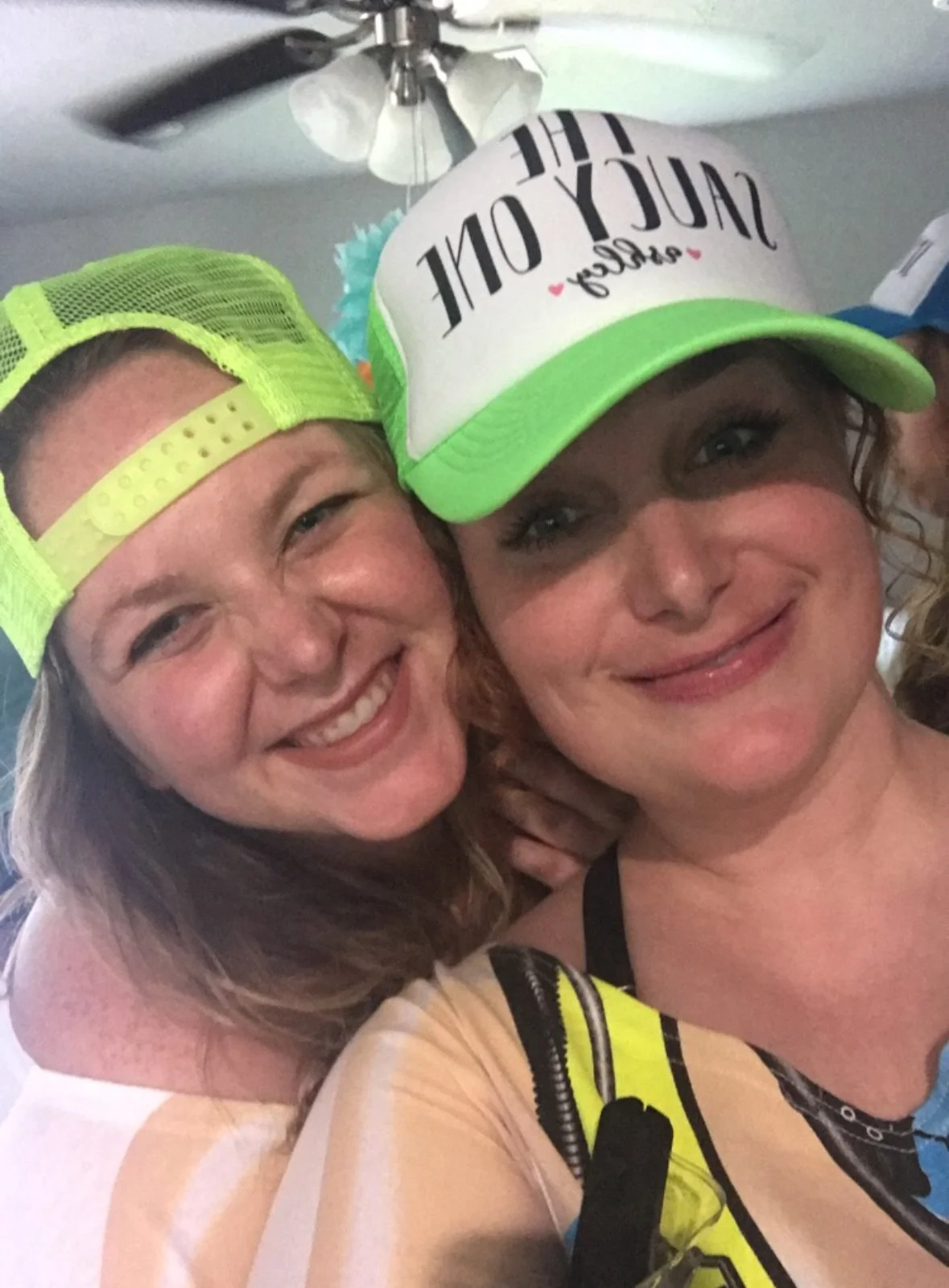 Two women smiling and taking a selfie indoors; one wearing a neon green cap and the other wearing a white cap with green accents that says 'Stay Out'.