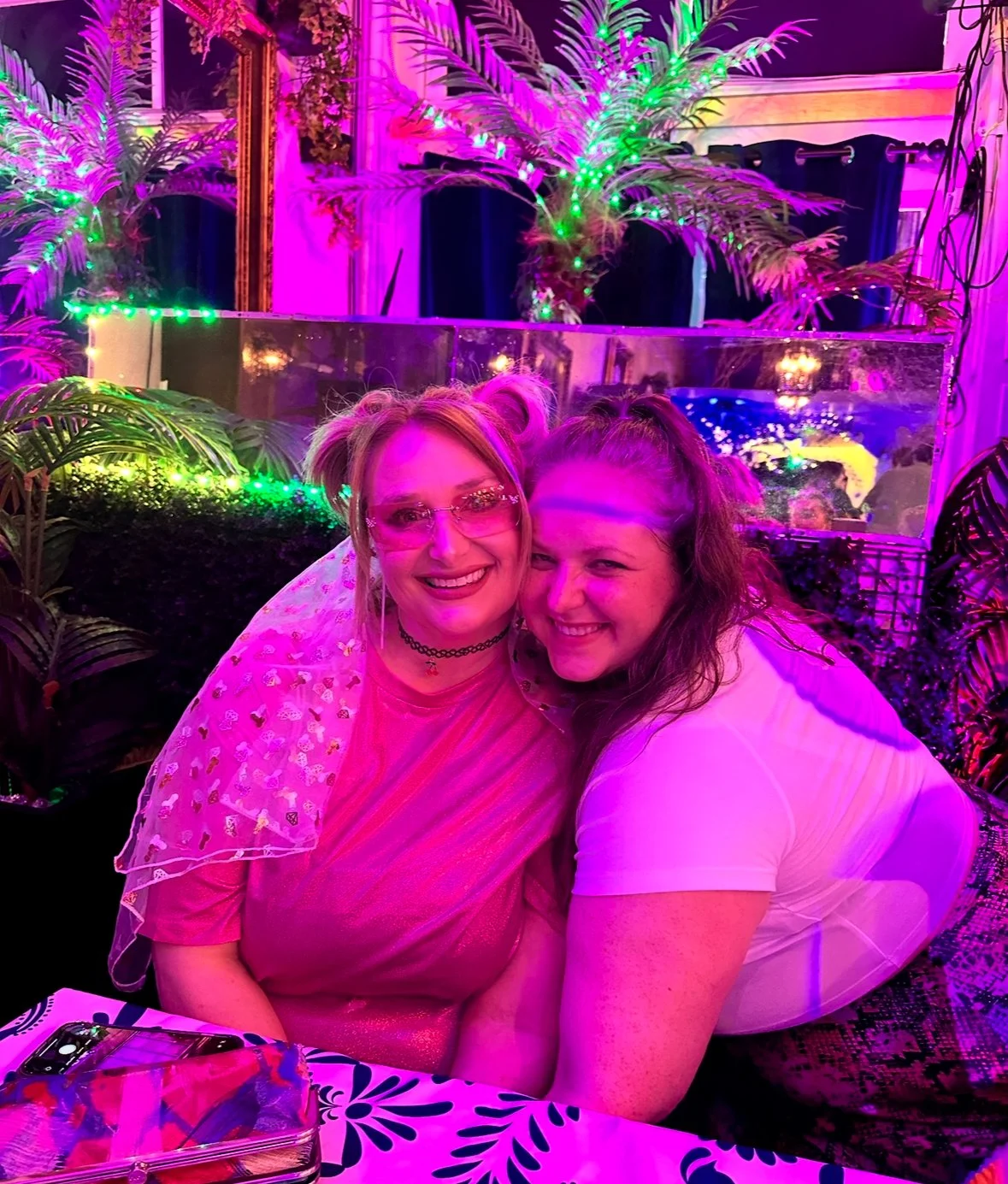 Two women are smiling and hugging at a colorful, neon-lit party with purple and green lighting, plants, and decorative elements in the background.