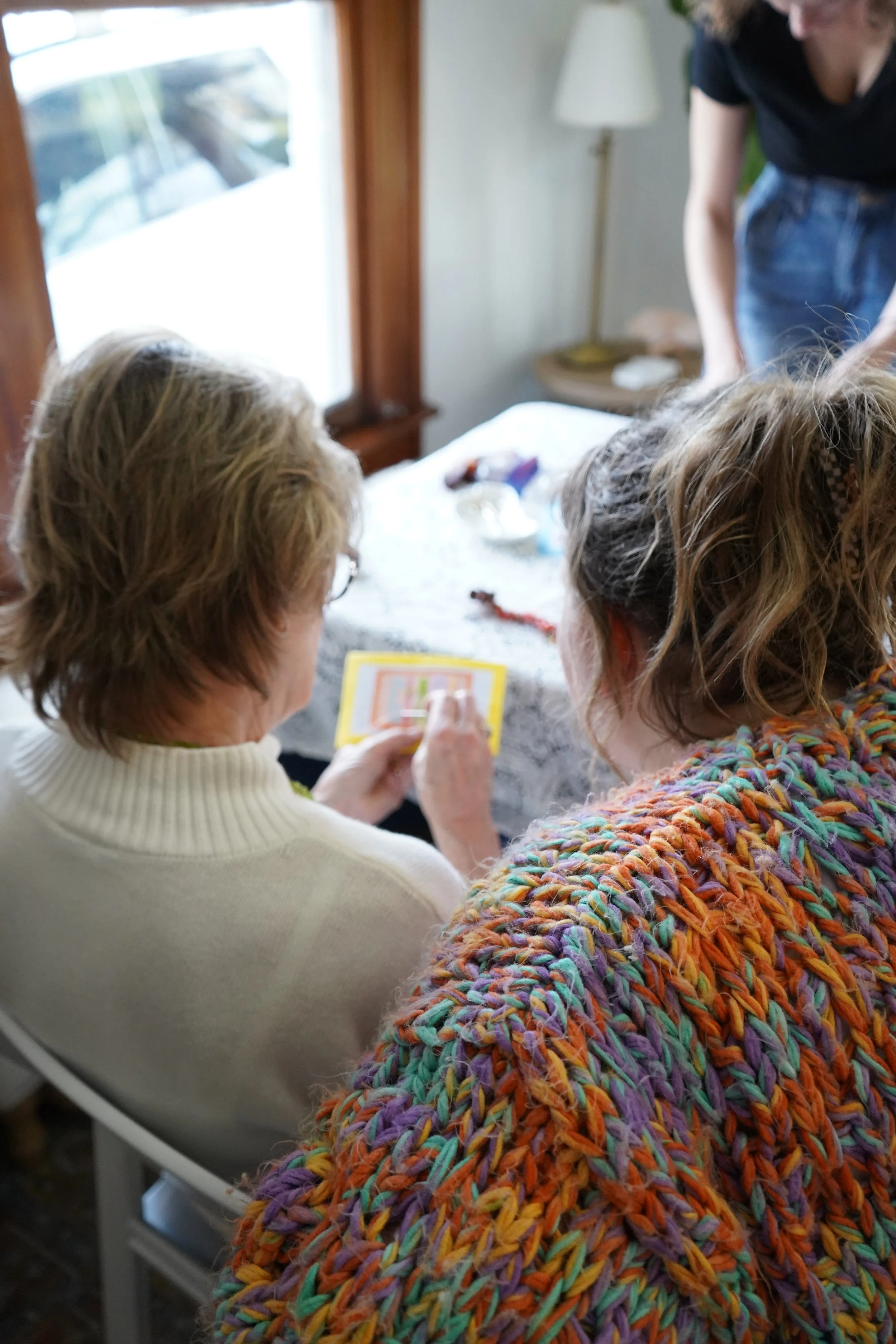 Two women sitting at a table with one holding a photo, while another woman stands nearby, in a cozy room with a window and lamps.