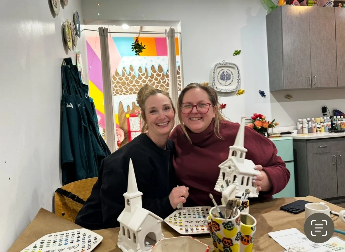 Two women sitting at a table, smiling, one holding a white ceramic church-shaped house, with arts and craft supplies on the table. The background has colorful decorations and a window with a creative mural.