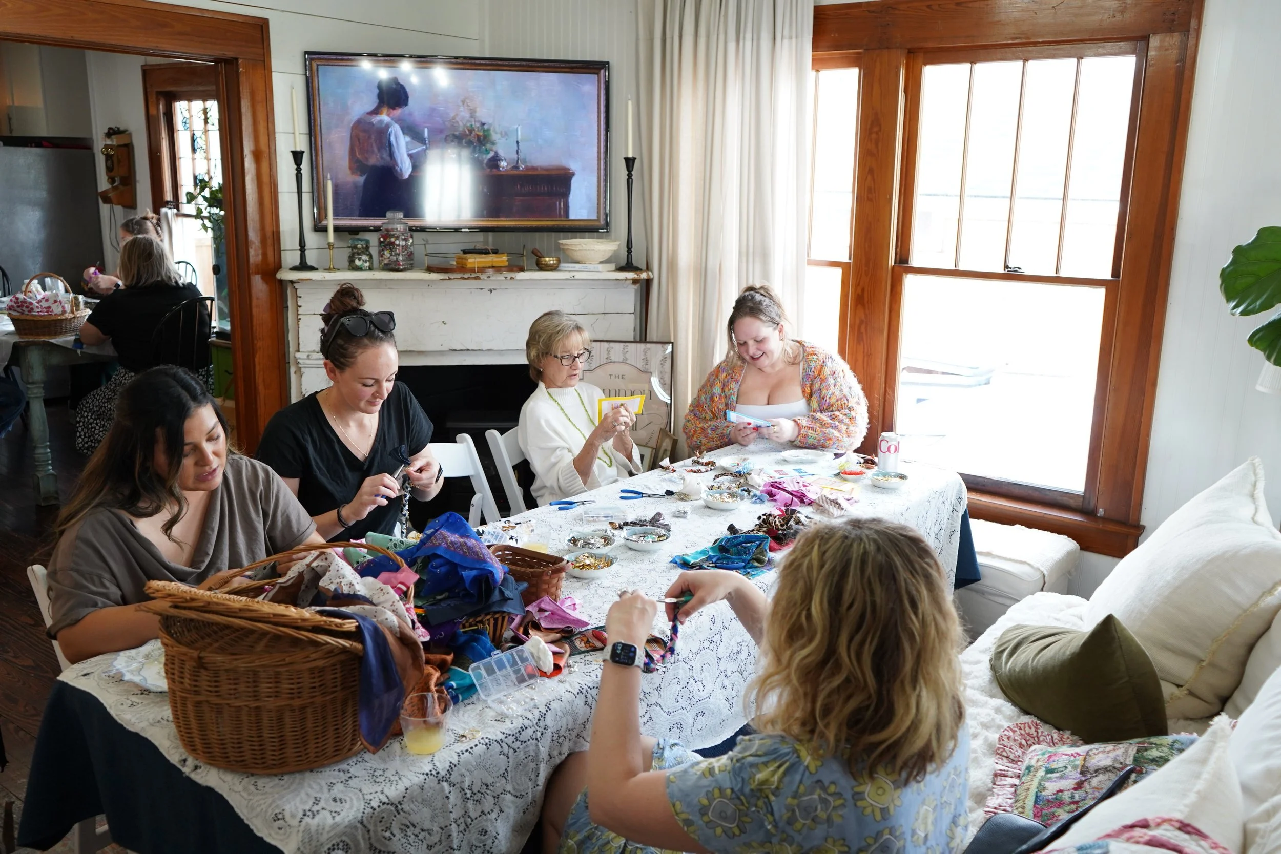 Group of women sitting around a table filled with craft supplies, making jewelry and chatting in a cozy, well-lit living room.