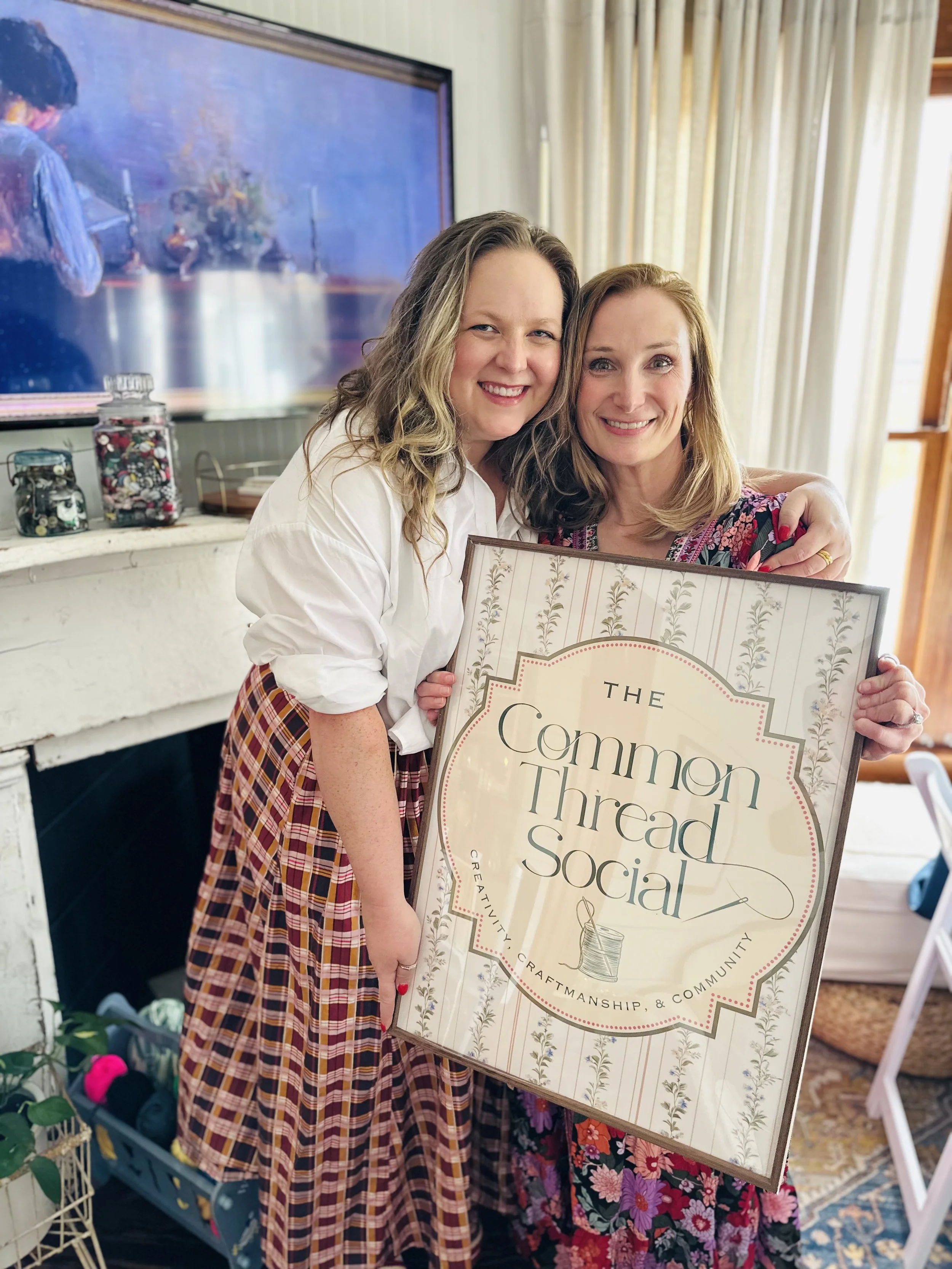 Two women smiling and hugging, holding a sign that says 'The Common Thread Social' in a cozy indoor setting with cream-colored curtains and a hearth where yarn and craft supplies are visible.