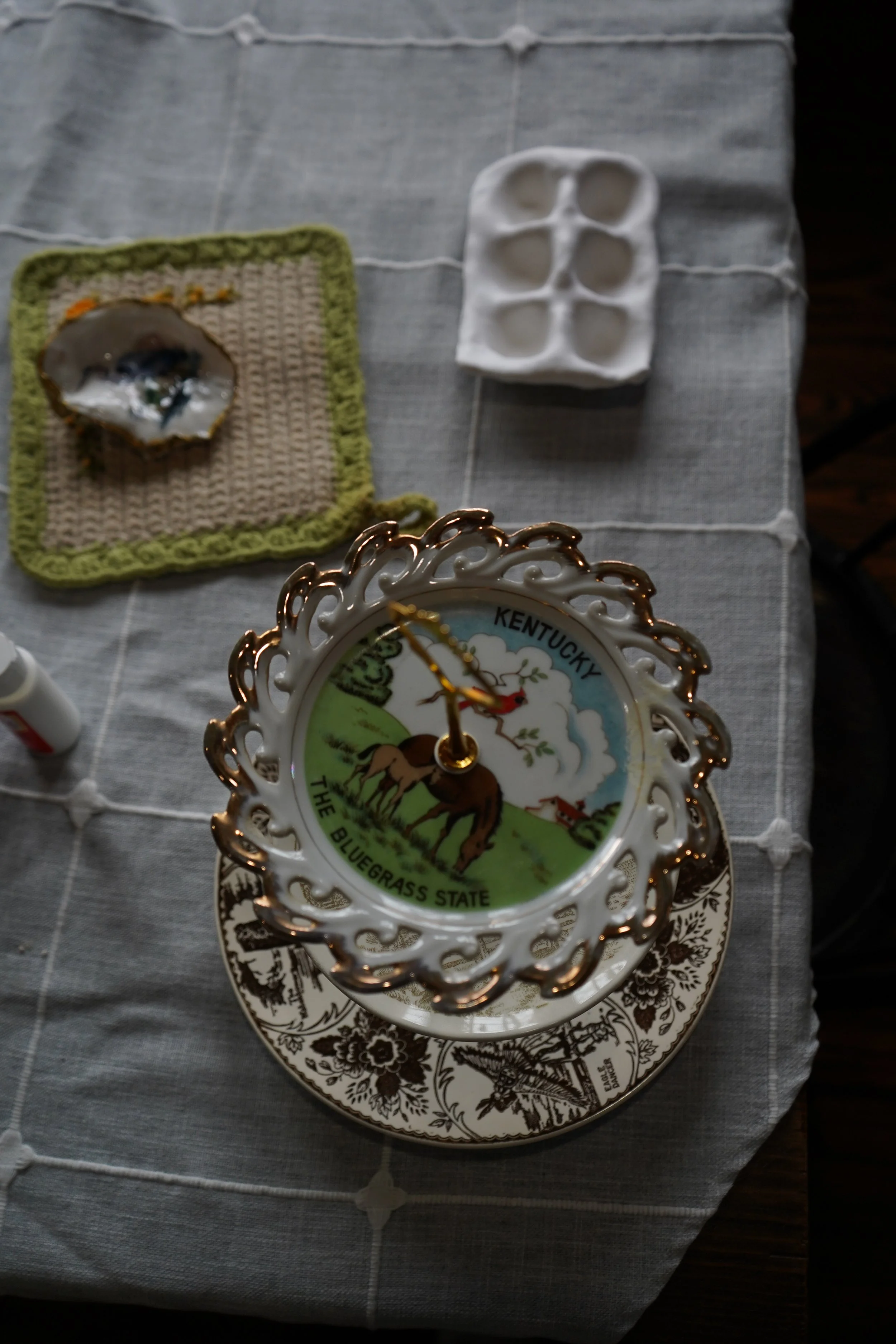 Decorative clock with Kentucky and Bluegrass State map, placed on a table with duck design, alongside a white egg carton, a small bowl with an oyster shell, and a crochet coaster with a painted design.