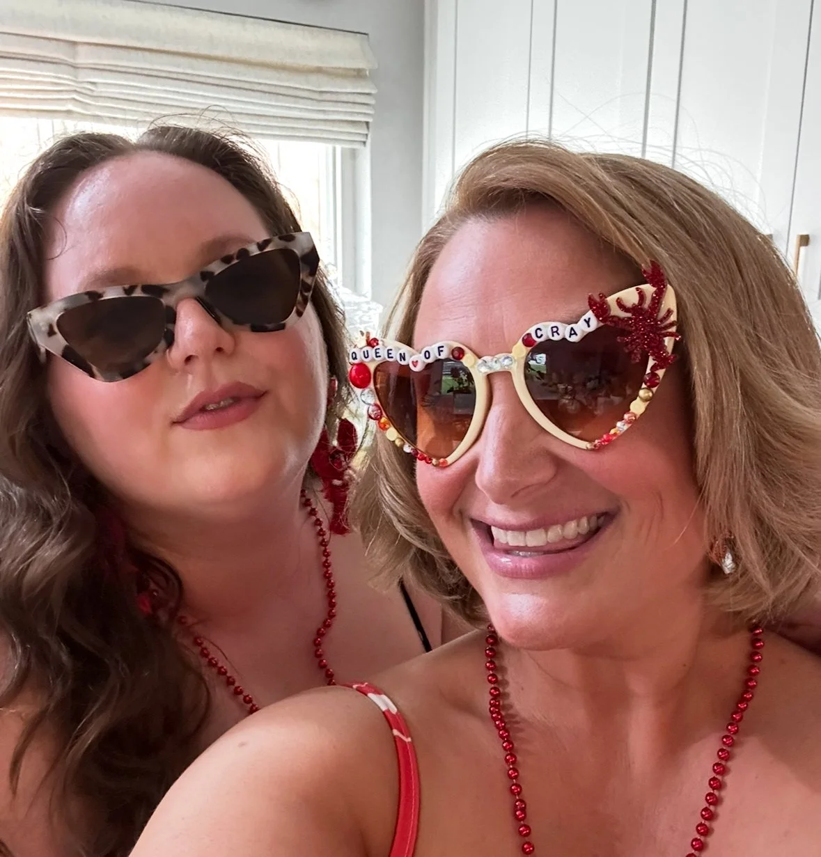 Two women wearing colorful, festive sunglasses, with the woman on the right smiling and the woman on the left looking at the camera.