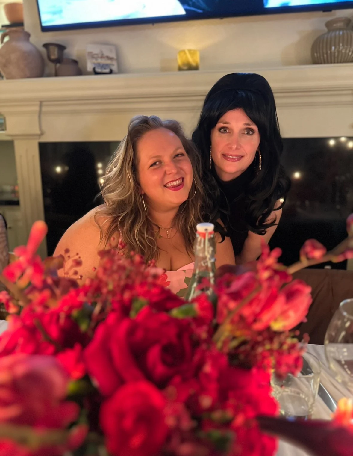 Two women smiling and posing for a photo at a gathering, with a bouquet of red flowers in the foreground and a fireplace behind them.