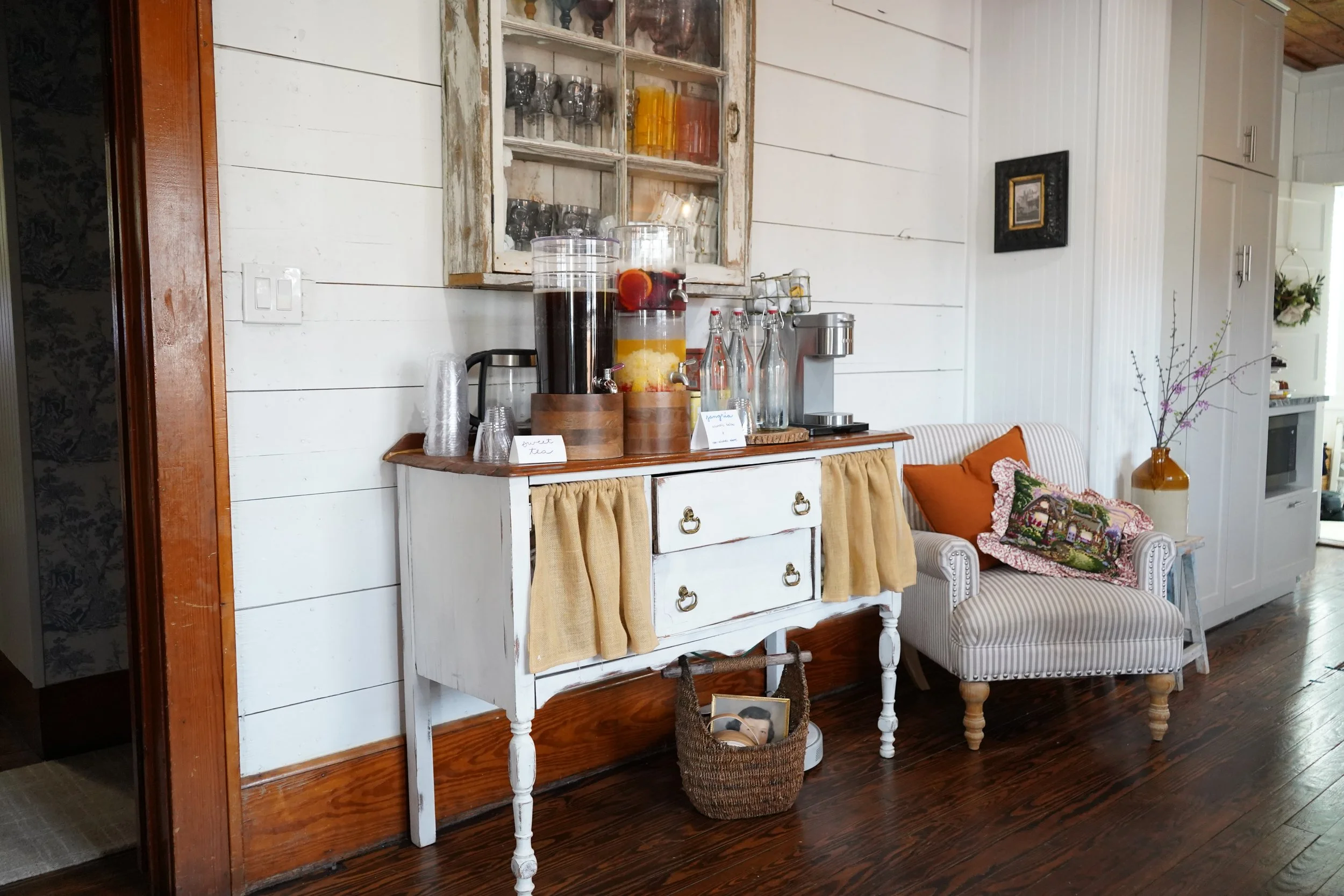 A rustic beverage station with two large dispensers of fruit-infused water on a distressed white sideboard, with glasses stacked nearby, set in a cozy room with wood-paneled walls and a striped armchair with pillows next to a vase with branches.