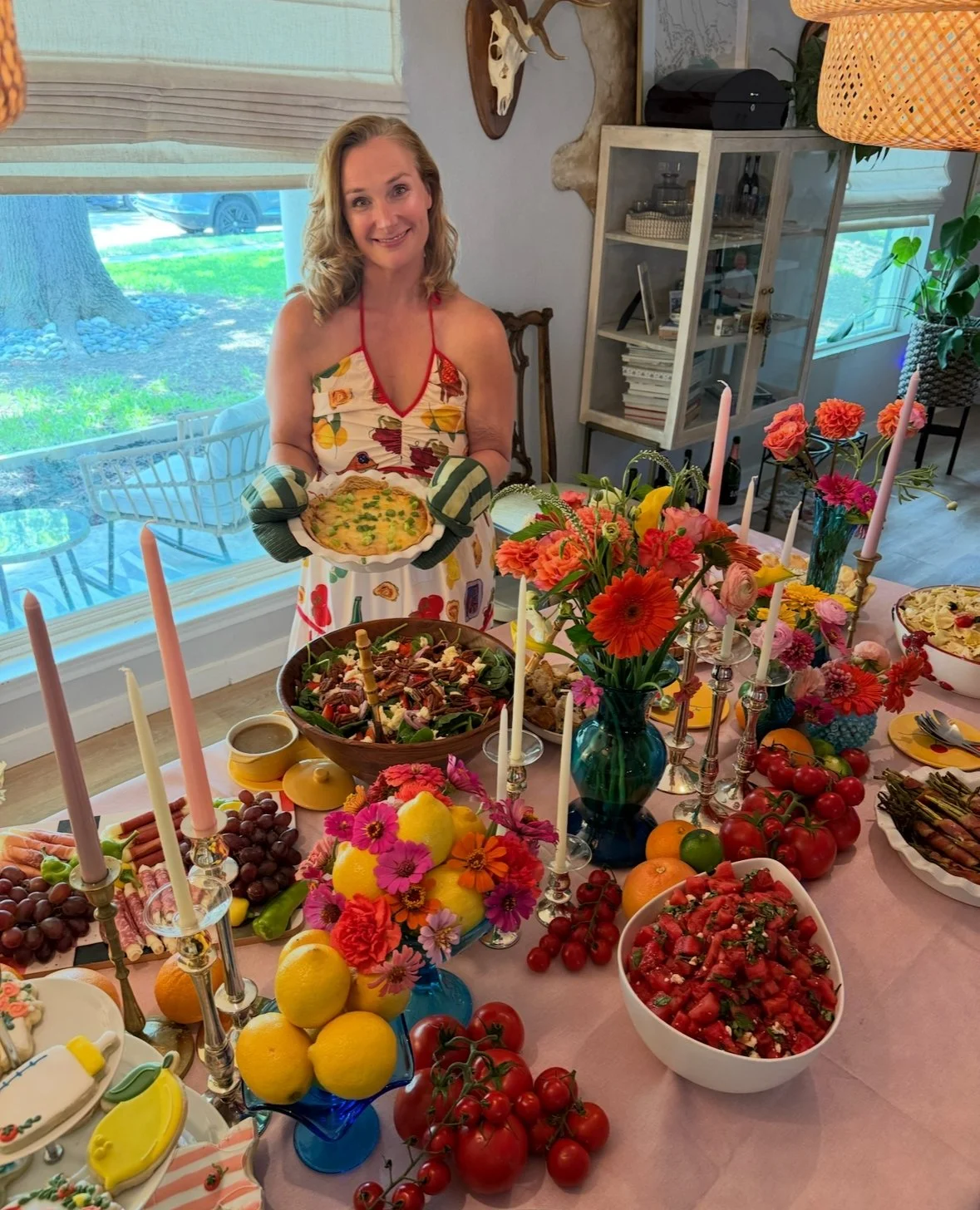 A woman in a colorful dress and oven mitts is holding a pie at a decorated dining table filled with flowers, fruits, salads, and candles, in a well-lit room with a large window showing green outdoor scenery.