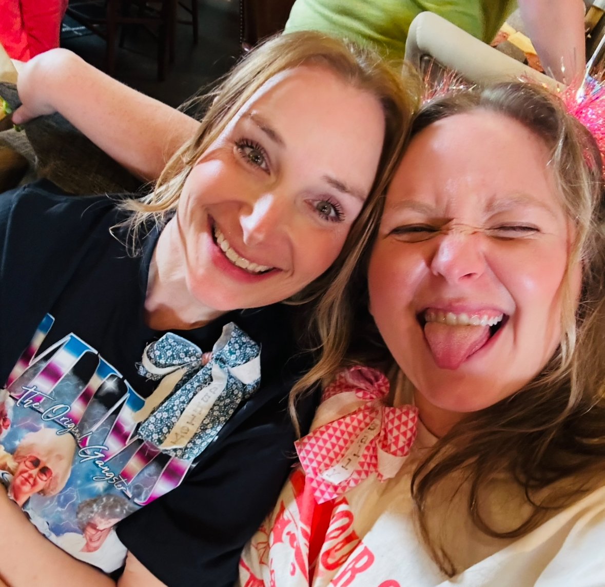 Two women taking a selfie together, both smiling and making playful faces, wearing colorful costumes and accessories, at a festive event.