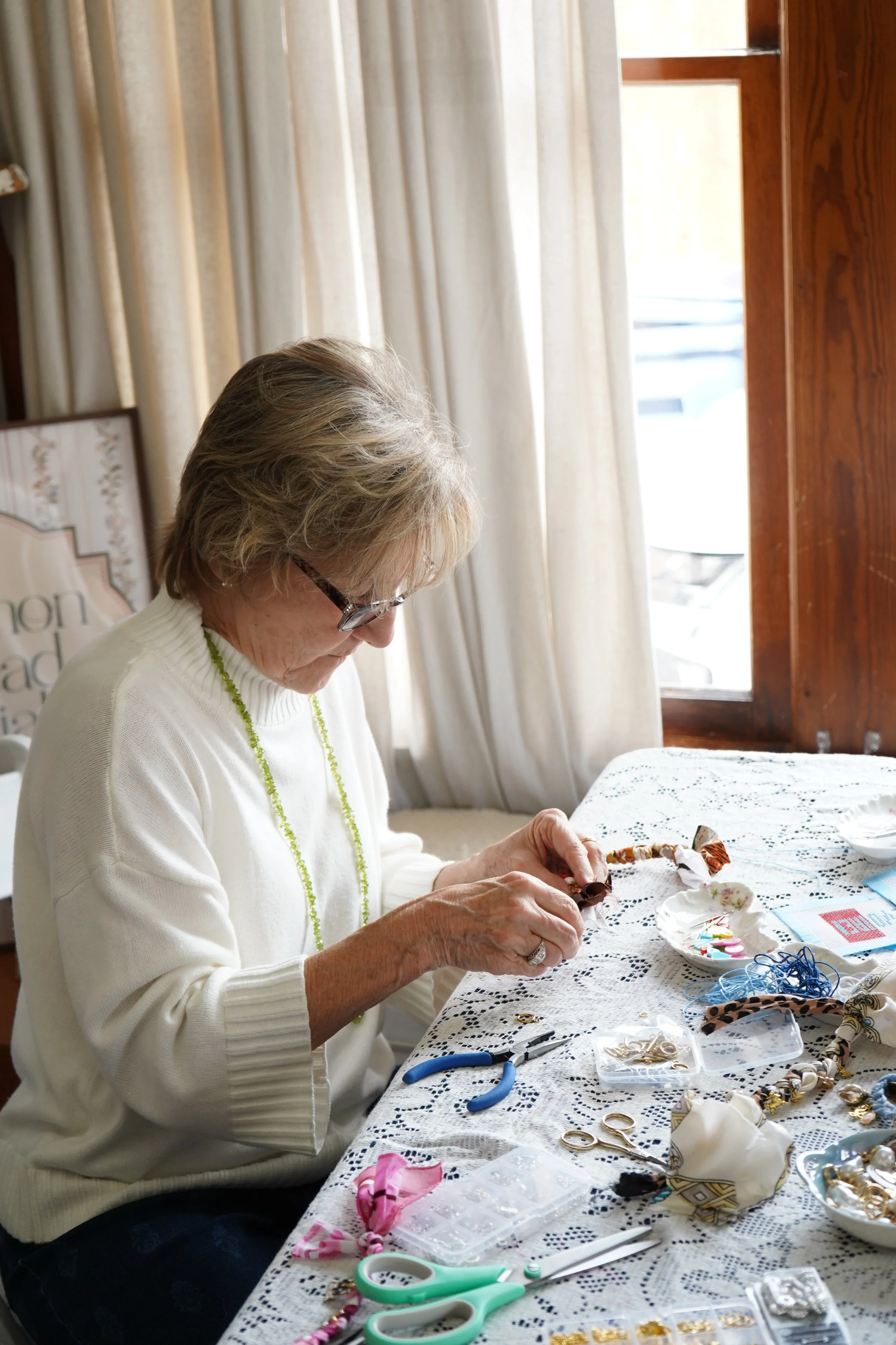 An elderly woman with glasses working on jewelry or craft projects at a table covered with craft supplies, including scissors, beads, and fabric, in a well-lit room with cream curtains and wooden window frames.
