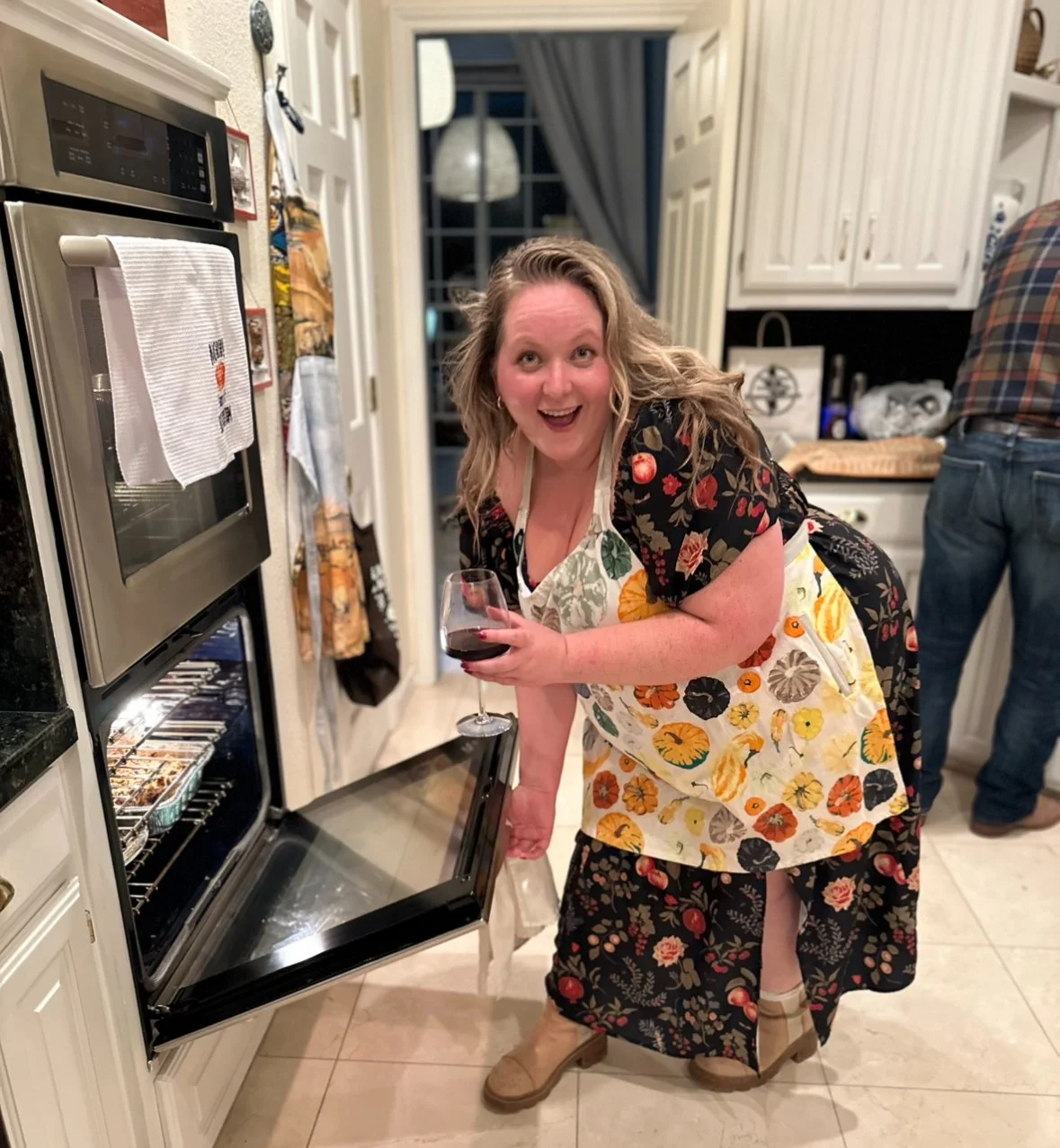 A woman with curly blonde hair wearing a black floral top and a yellow floral apron, holding a glass of red wine, smiling cheerfully in a home kitchen while leaning over an open oven door. Another person is partially visible in the background.
