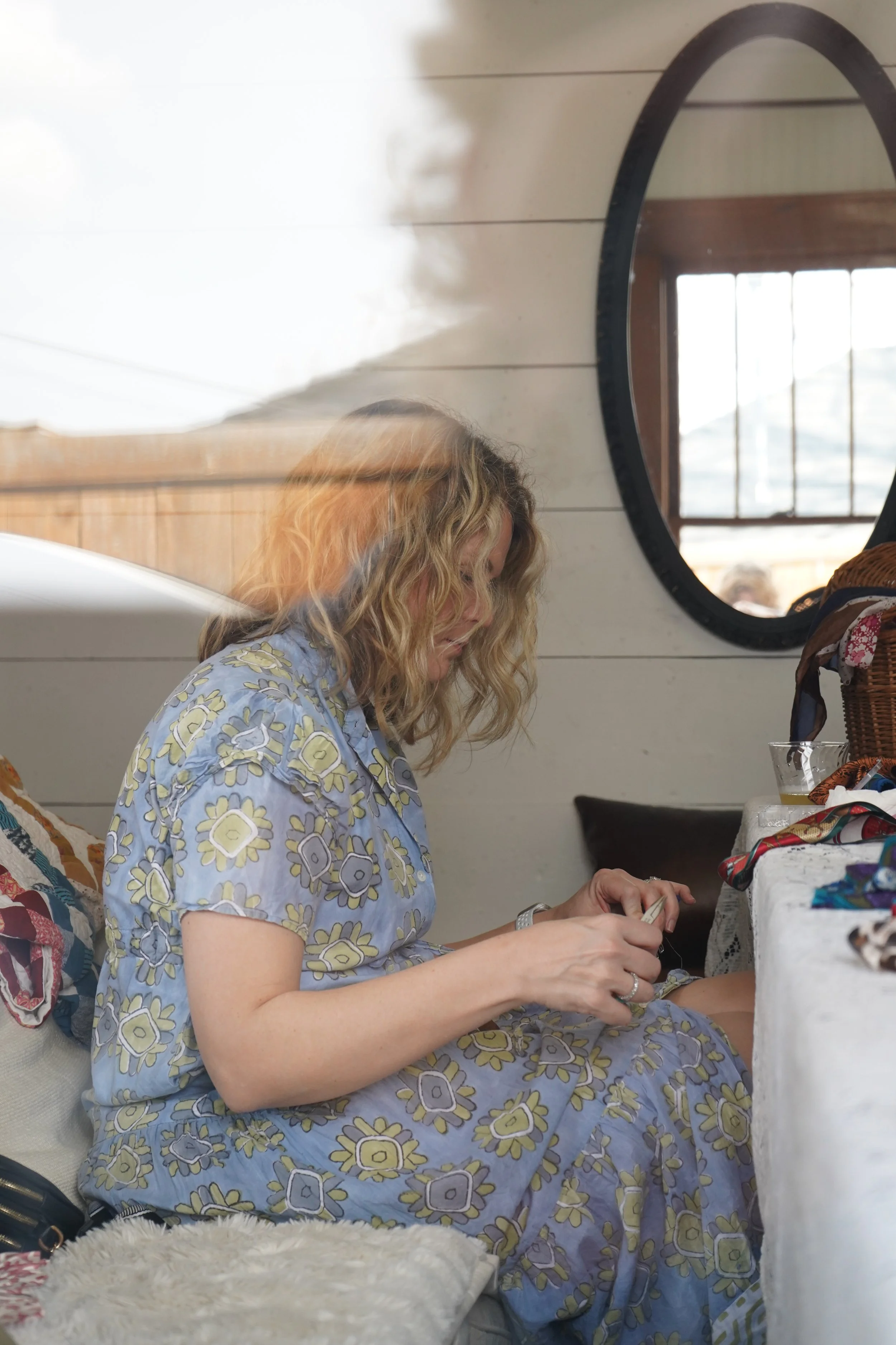 A woman with curly blonde hair sitting at a table, wearing a blue floral dress, and looking at her hands. The table has various items on it, and a window with a circular mirror reflecting the outdoors is in the background.