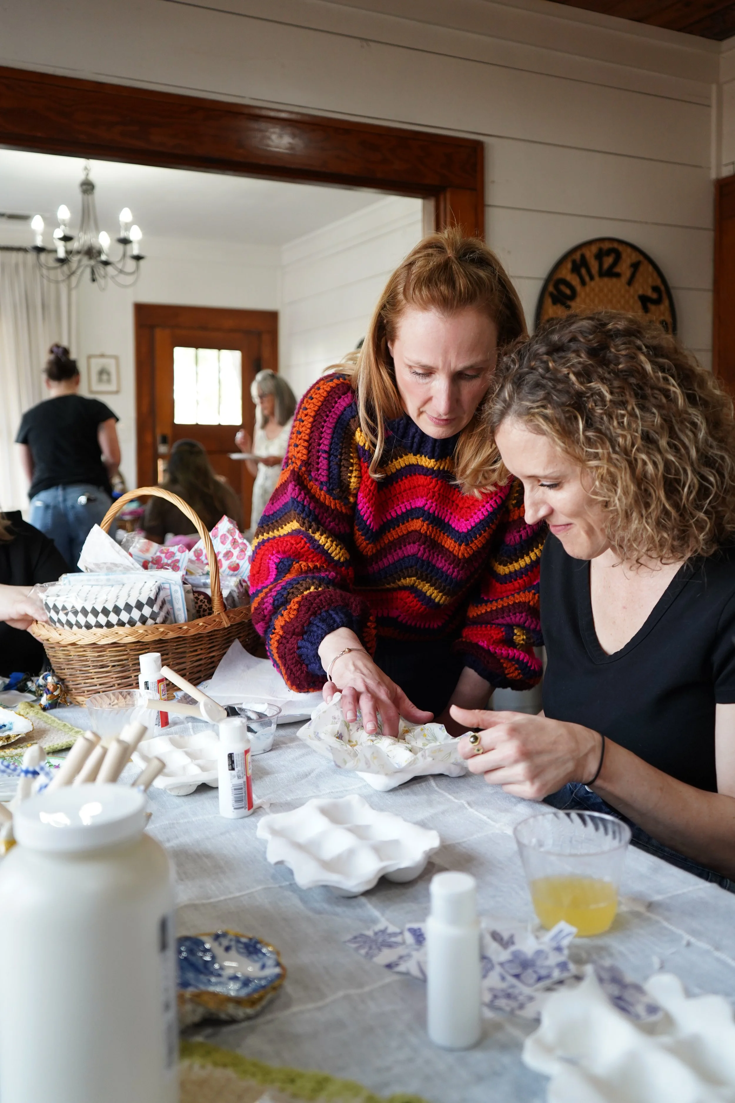 Two women are working together on a craft project at a table, surrounded by craft supplies including glue, paint, and paper, in a cozy room with a large clock on the wall and other people in the background.
