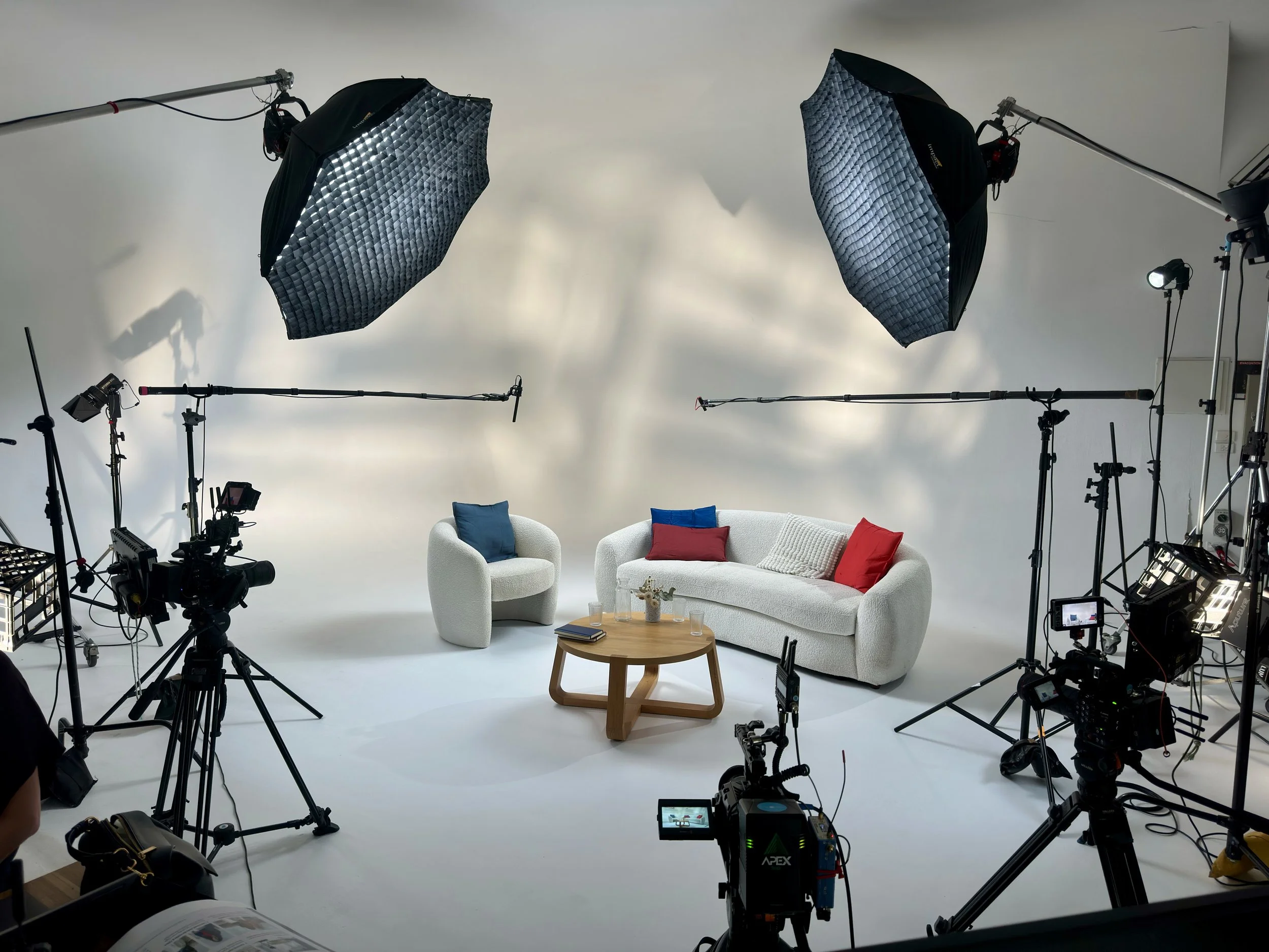 A professional video or photo studio setup with a white sofa, a white armchair, a small wooden table with glasses and a book, surrounded by large studio lights with umbrellas, cameras, and other equipment for filming or photography.