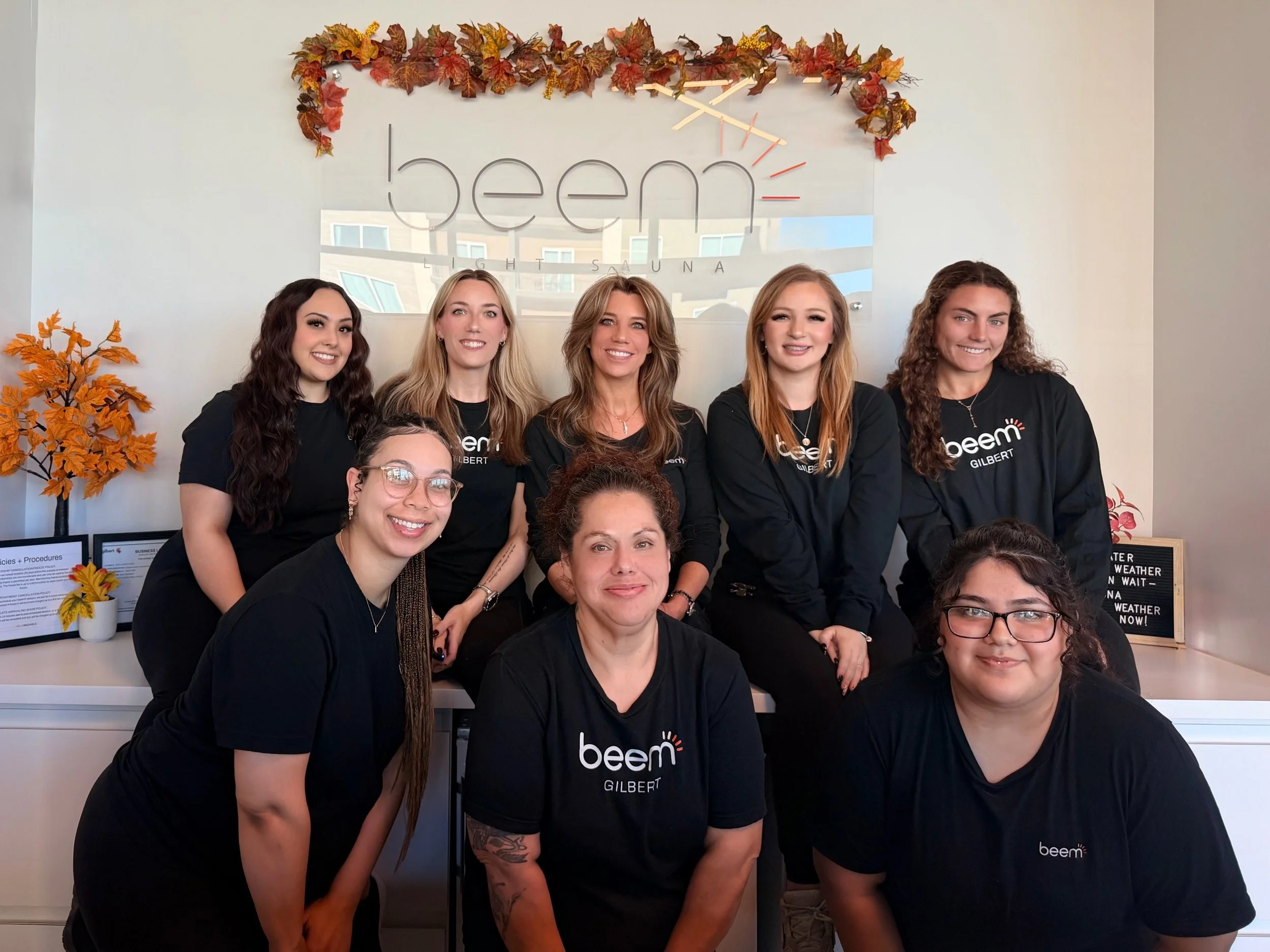 A group of women wearing matching black t-shirts with the word 'beem' on it, posing together indoors in front of a decorative autumn-themed wall with fall leaves and a sign. They are smiling and standing close together, with some women sitting and others standing.