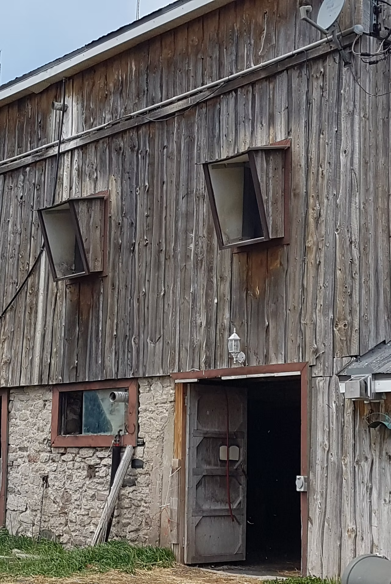 The exterior of a rustic barn with weathered wooden siding and a stone foundation. Two open windows with tilted panes, a small window with a pipe, an open door, a lantern above the door, and various utility wires and equipment are visible.