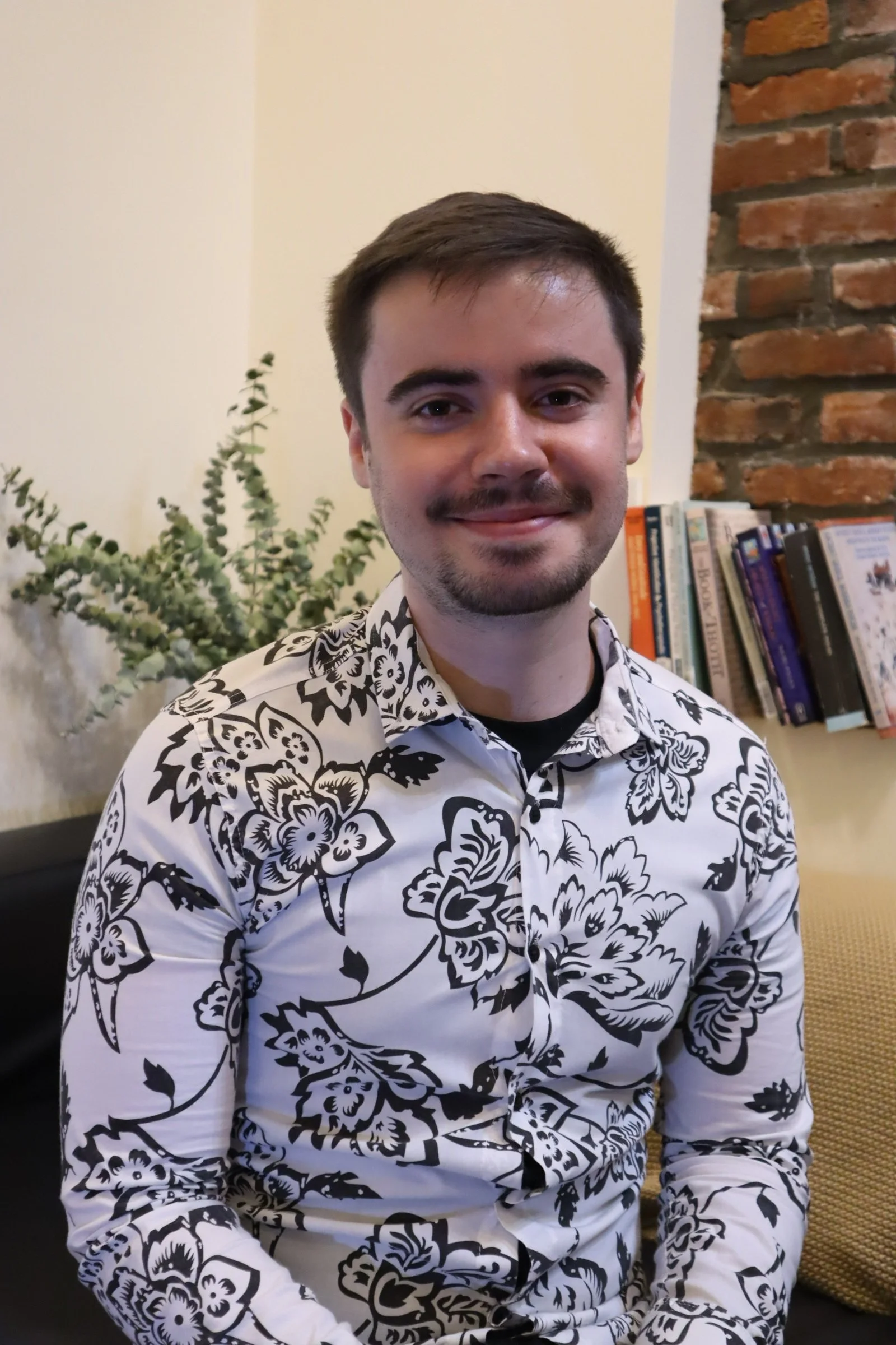 A young man with short dark hair and a light beard, smiling, wearing a black and white floral patterned shirt, sitting indoors with a bookshelf and a leafy plant in the background.