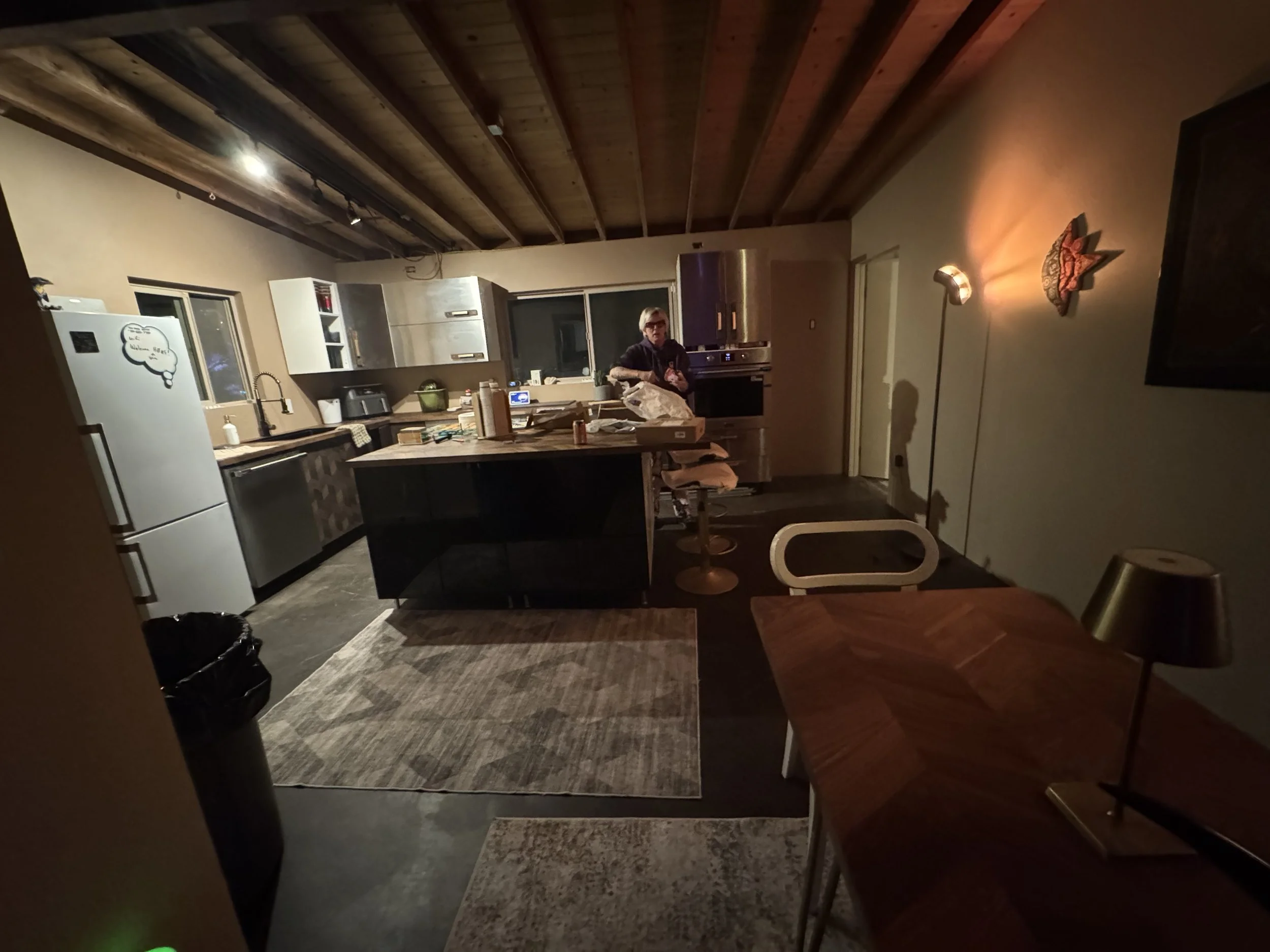 A person standing in a kitchen with modern appliances and wooden ceiling, kitchen island in the center, and a dining table with chairs nearby. Indoor lighting creates a cozy atmosphere.