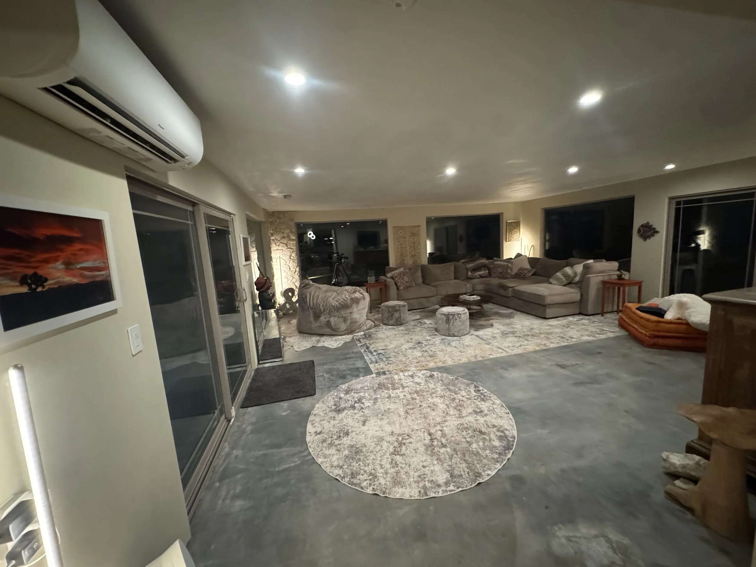 A spacious living room with a large beige sectional sofa, a patterned area rug, and various chairs and tables. The room features large windows, glass sliding doors, and a stone accent wall, with modern lighting mounted on the ceiling.
