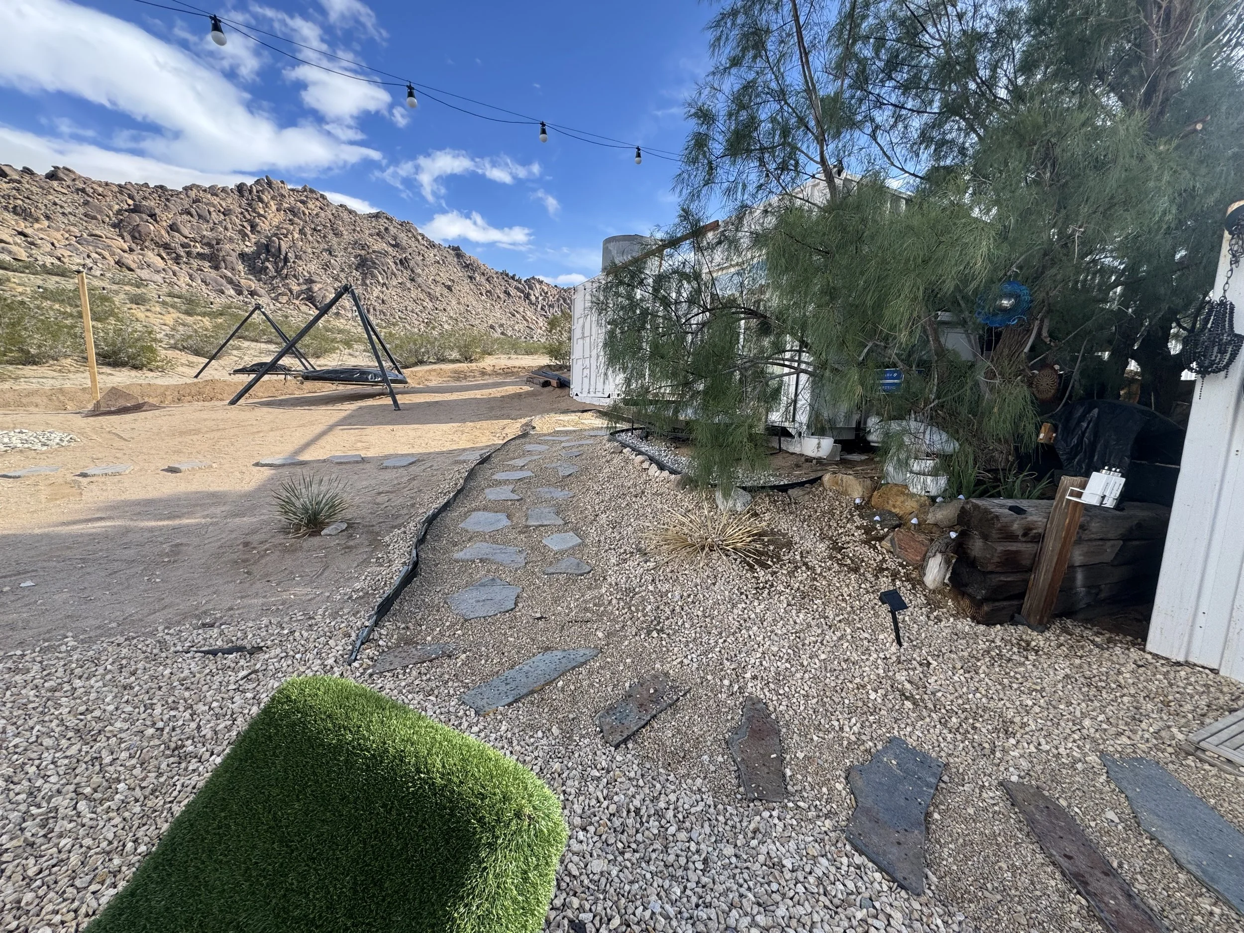 Desert yard with gravel ground, stepping stones, a green grass patch, a large tree, a slide, and a mountain backdrop under a blue sky with clouds.