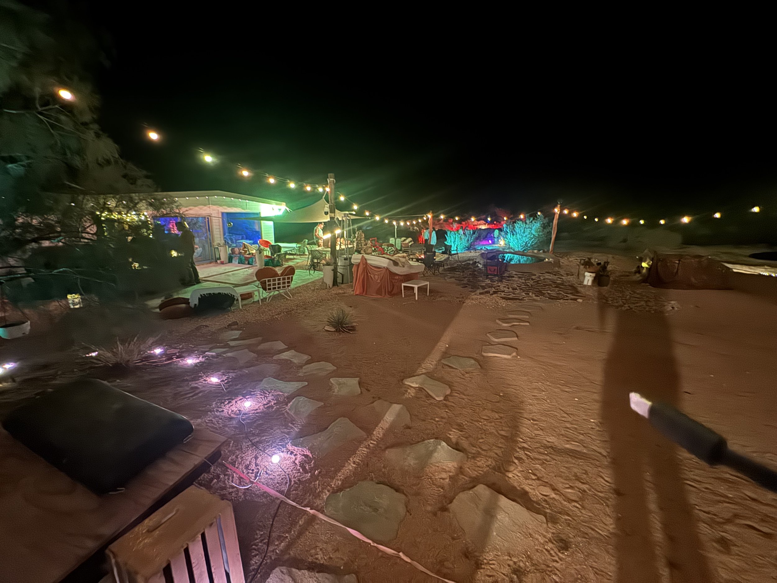 Nighttime outdoor party scene with colorful string lights, a house with illuminated section, chairs, tables, and trees decorated with lights, set on a sandy terrain.