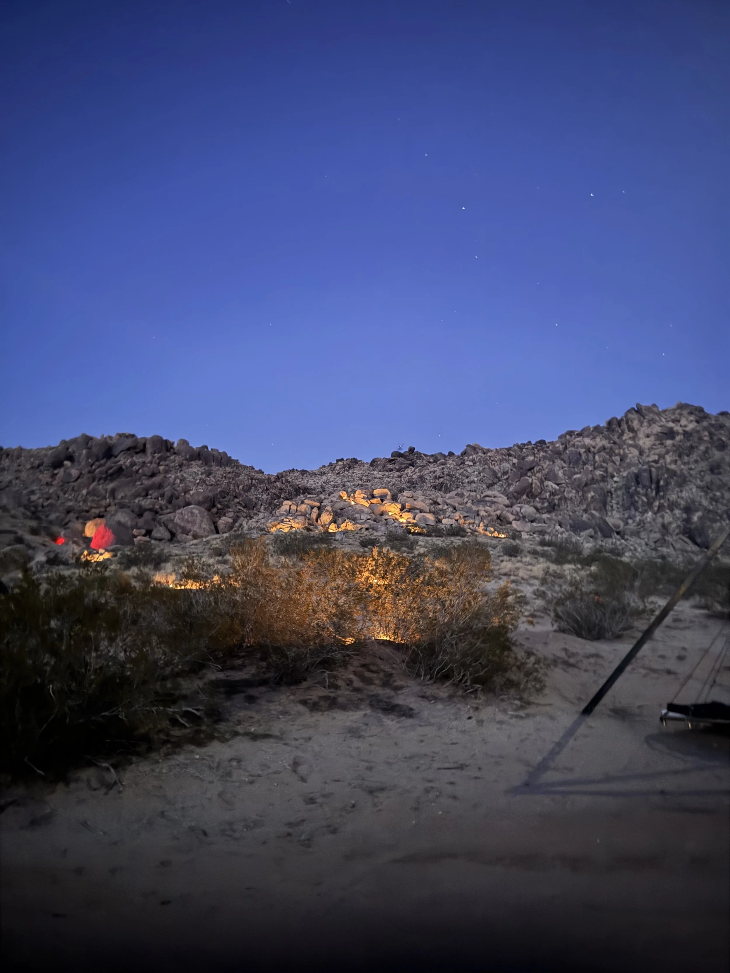 Nighttime desert scene with rocky hills illuminated by warm lights, sandy ground with sparse bushes, and a clear blue sky with visible stars.