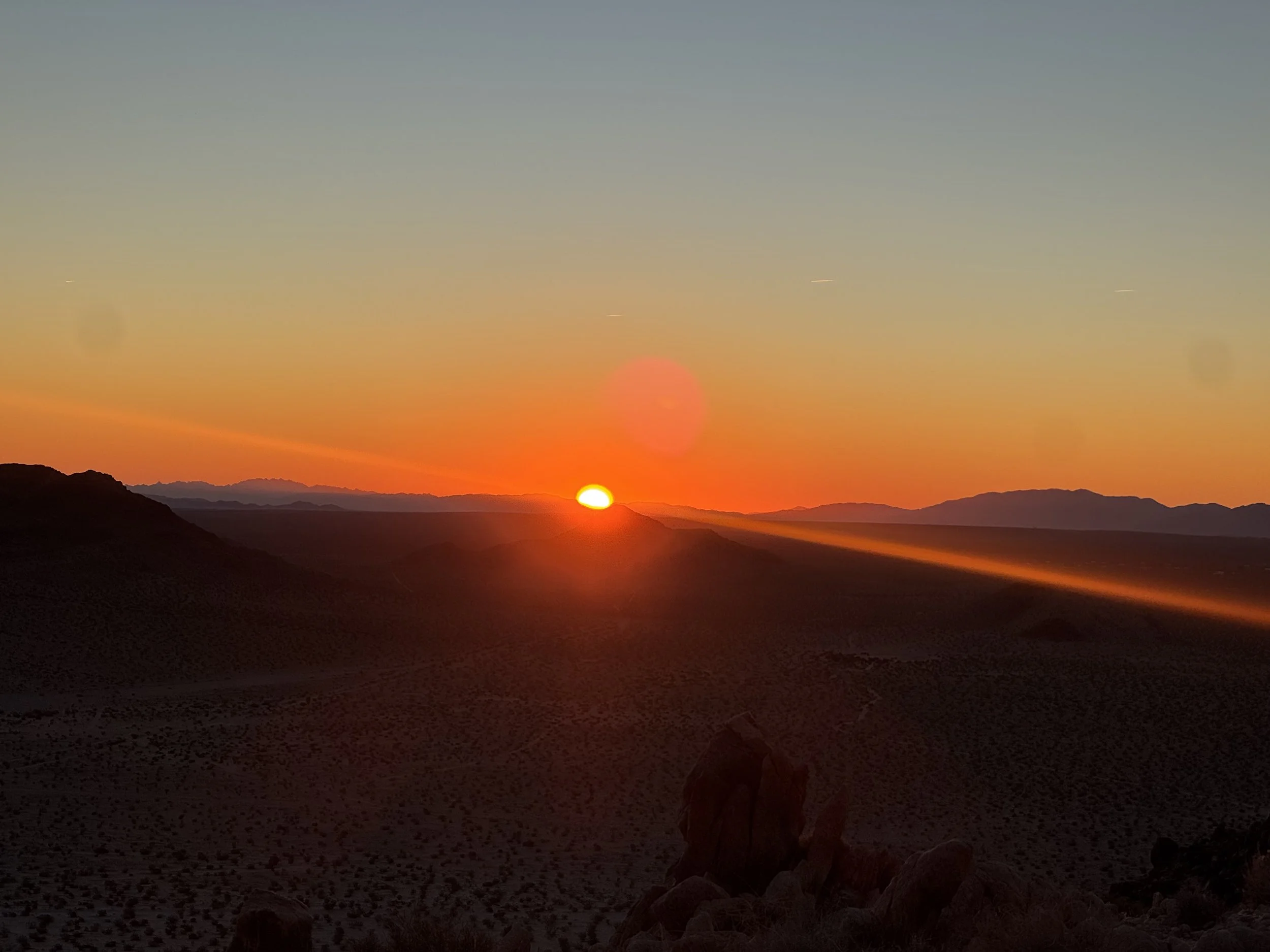 Sunset over a desert landscape with silhouettes of hills and mountains in the distance.