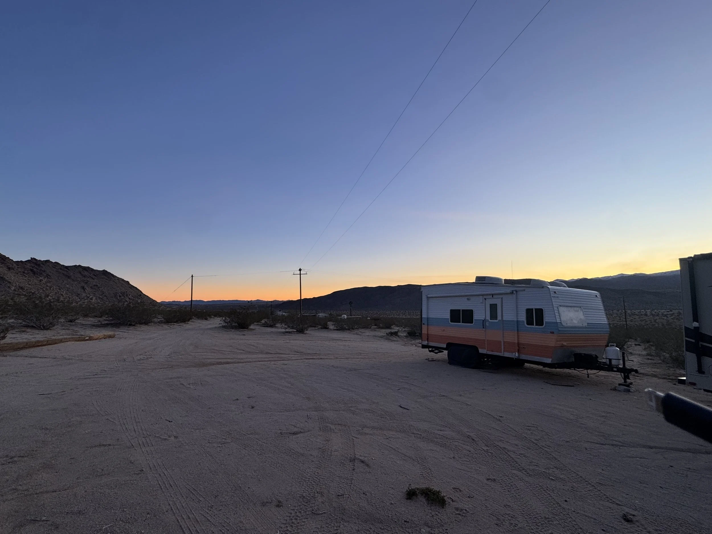 A vintage camper parked on a dirt road in a desert landscape during sunset, with mountains in the background and a clear sky.