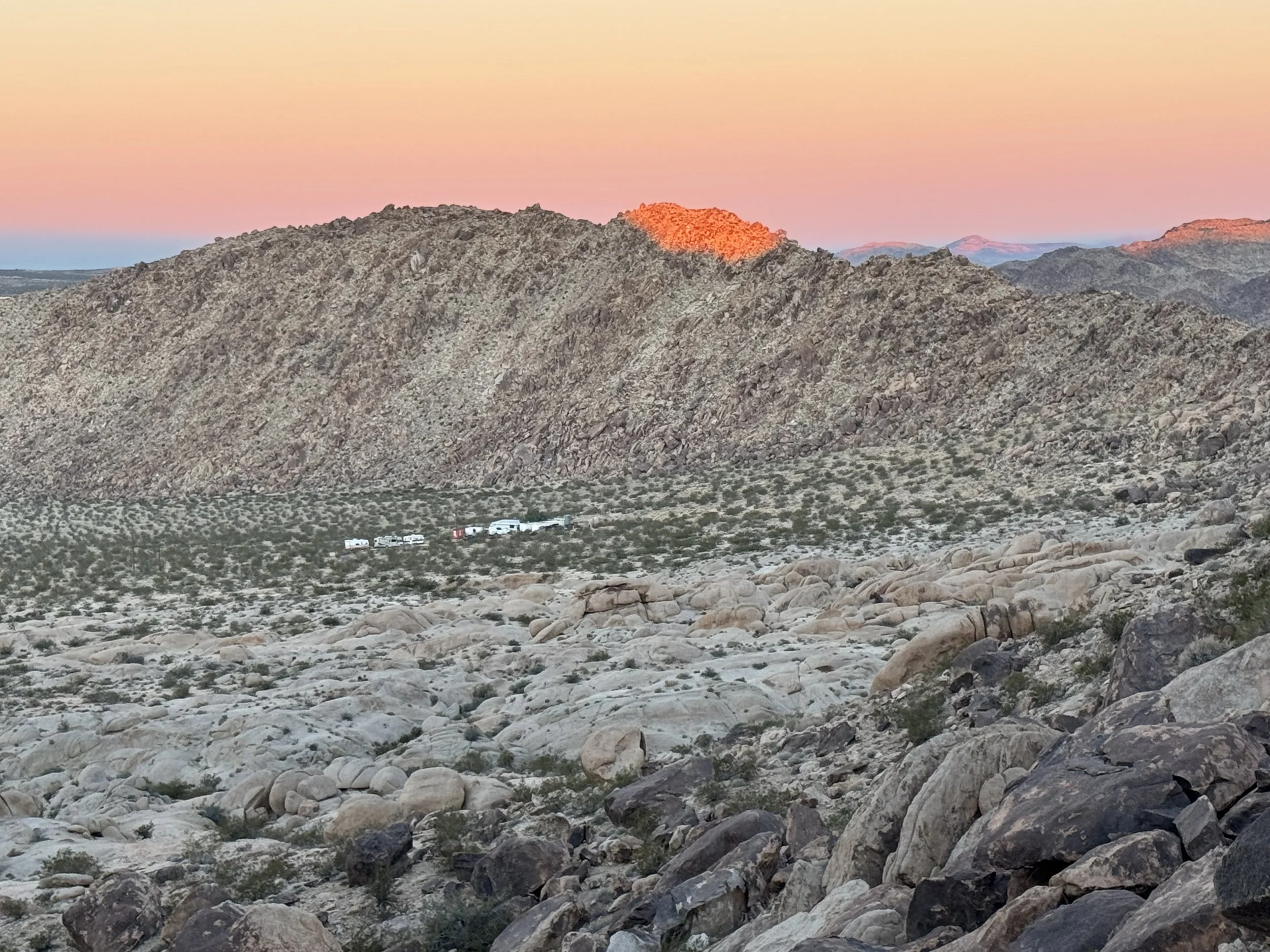 A desert landscape with rocky terrain and sparse vegetation, a mountain range in the background, and a colorful sky at sunset with pink and orange hues.