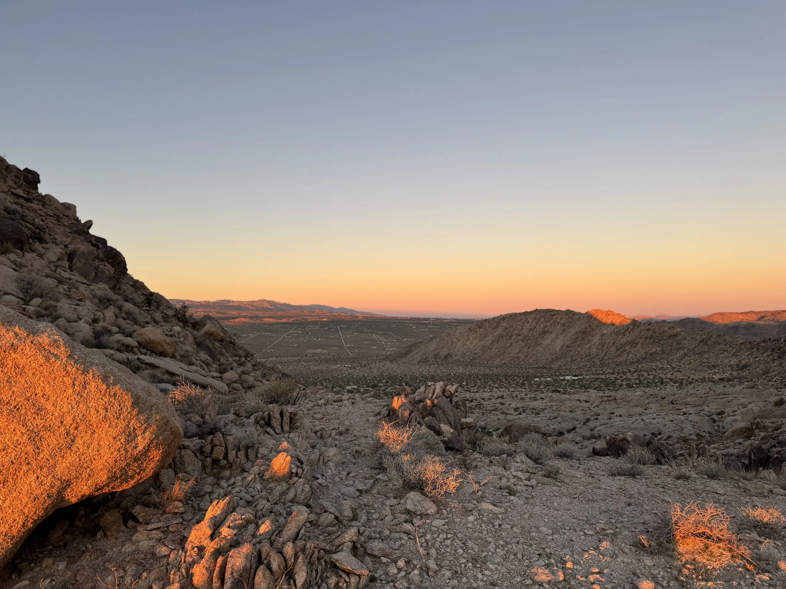 Desert landscape at sunset with rocky terrain, sparse vegetation, and distant hills under a clear blue sky.