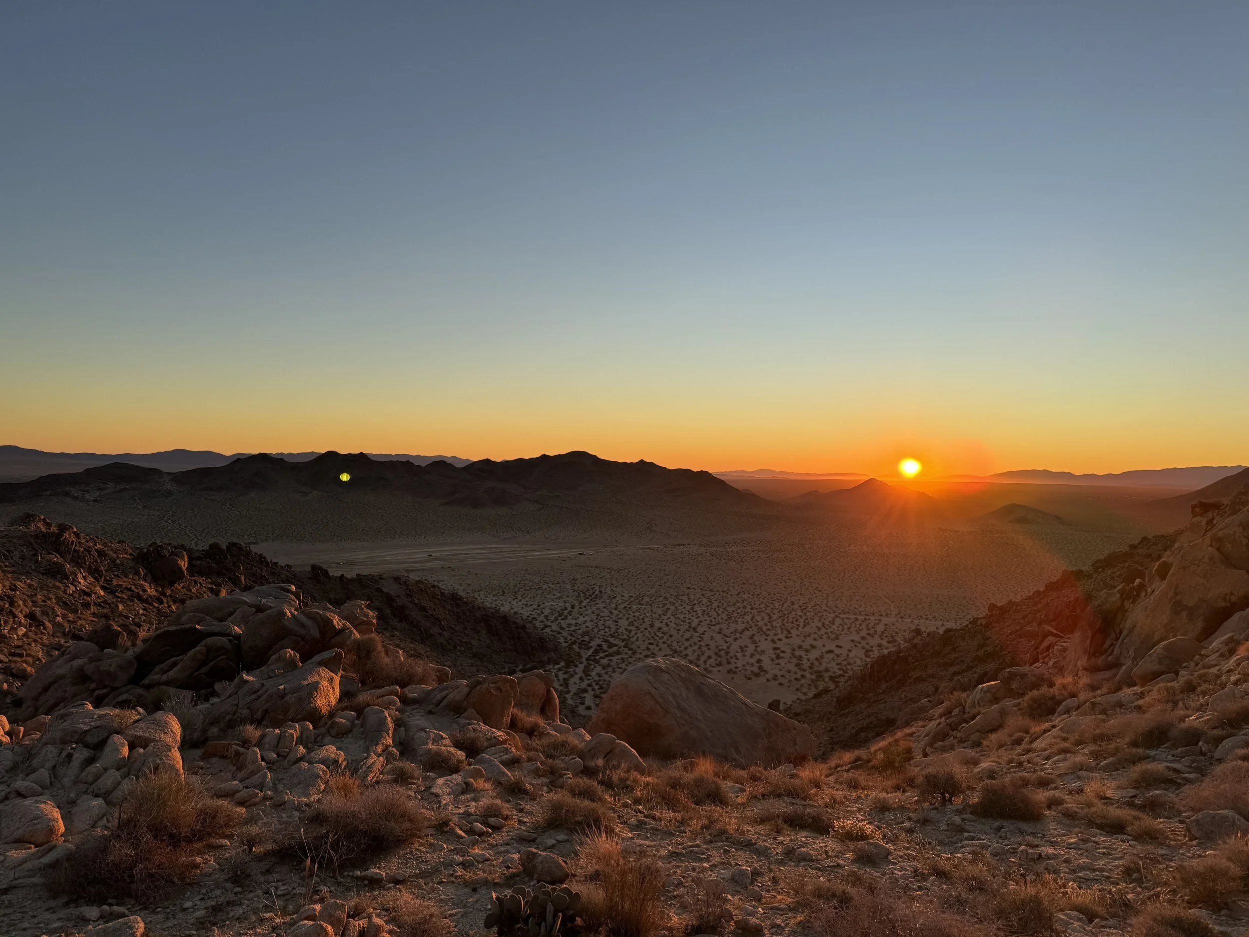 Desert landscape during sunset with rocky terrain, sparse vegetation, distant mountains, and a bright sun low on the horizon.