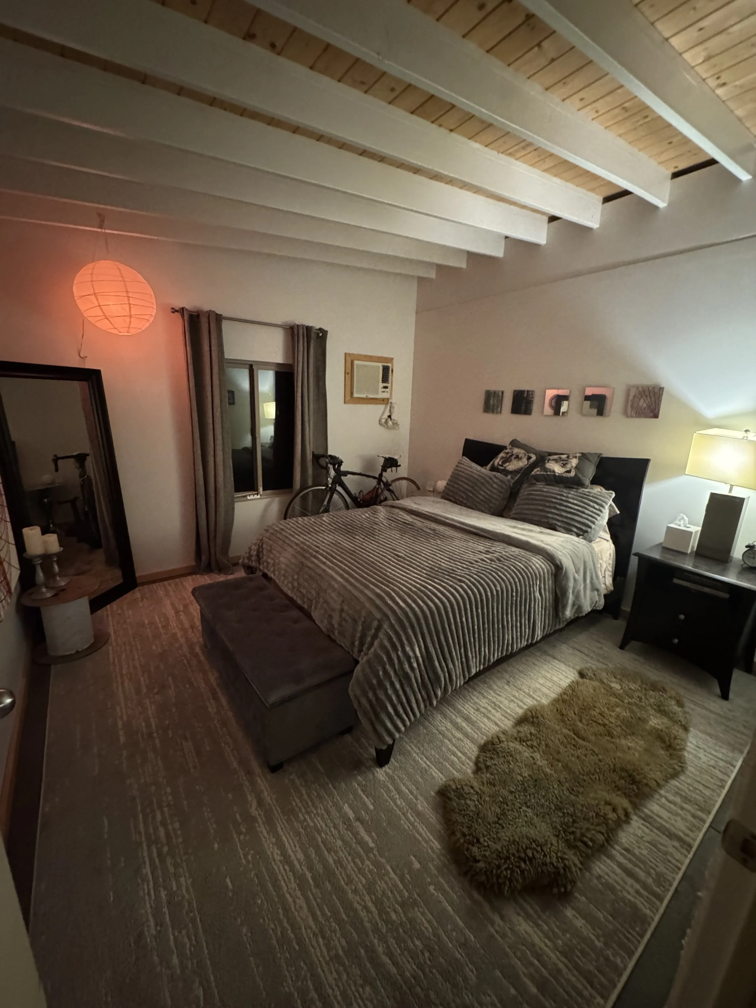 A cozy bedroom with a neatly made bed covered with a striped blanket, several pillows, and a headboard. There is a nightstand with a lamp and tissue box beside the bed, a textured rug on the floor, and a small stuffed animal at the foot of the bed. A