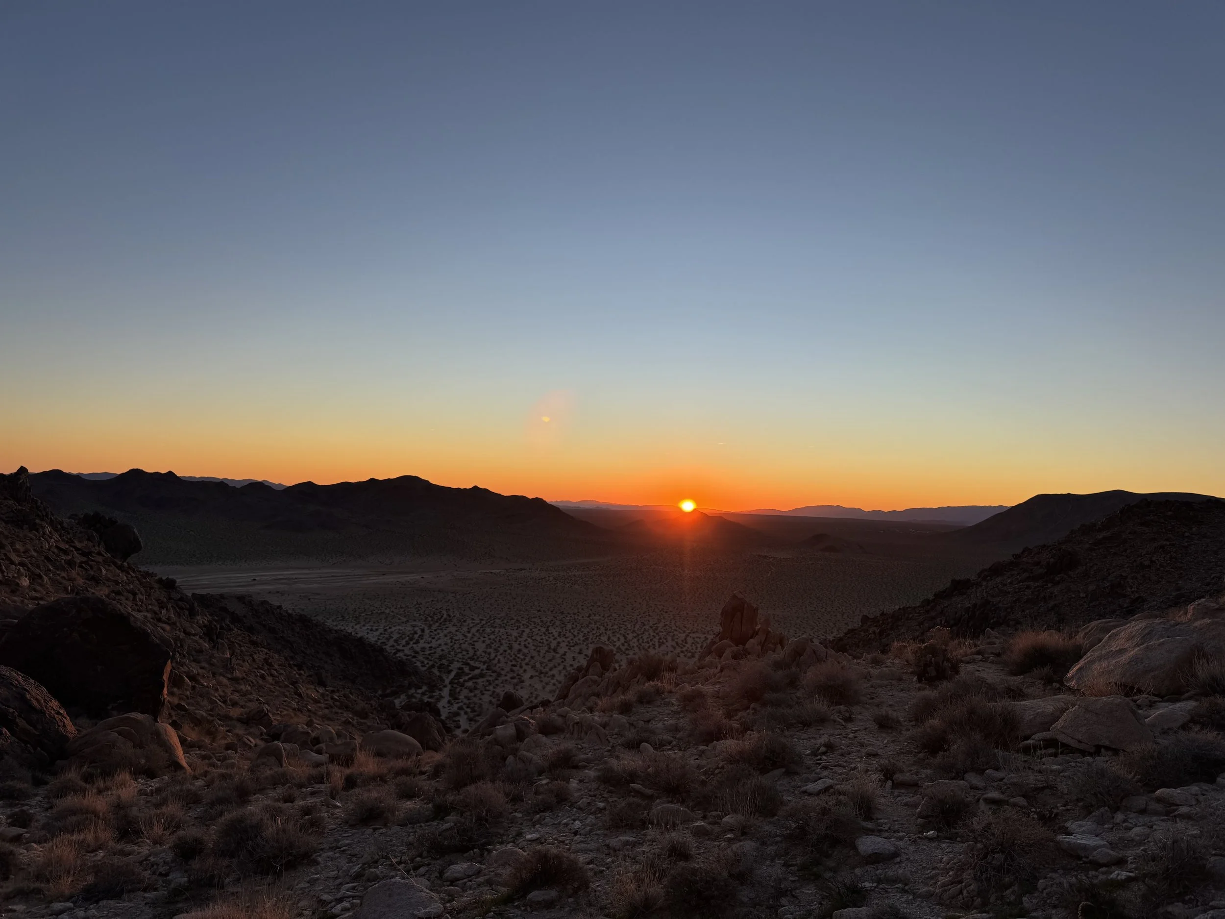 Desert landscape during sunset with rocky terrain, sparse bushes, distant mountains, and a setting sun on the horizon.