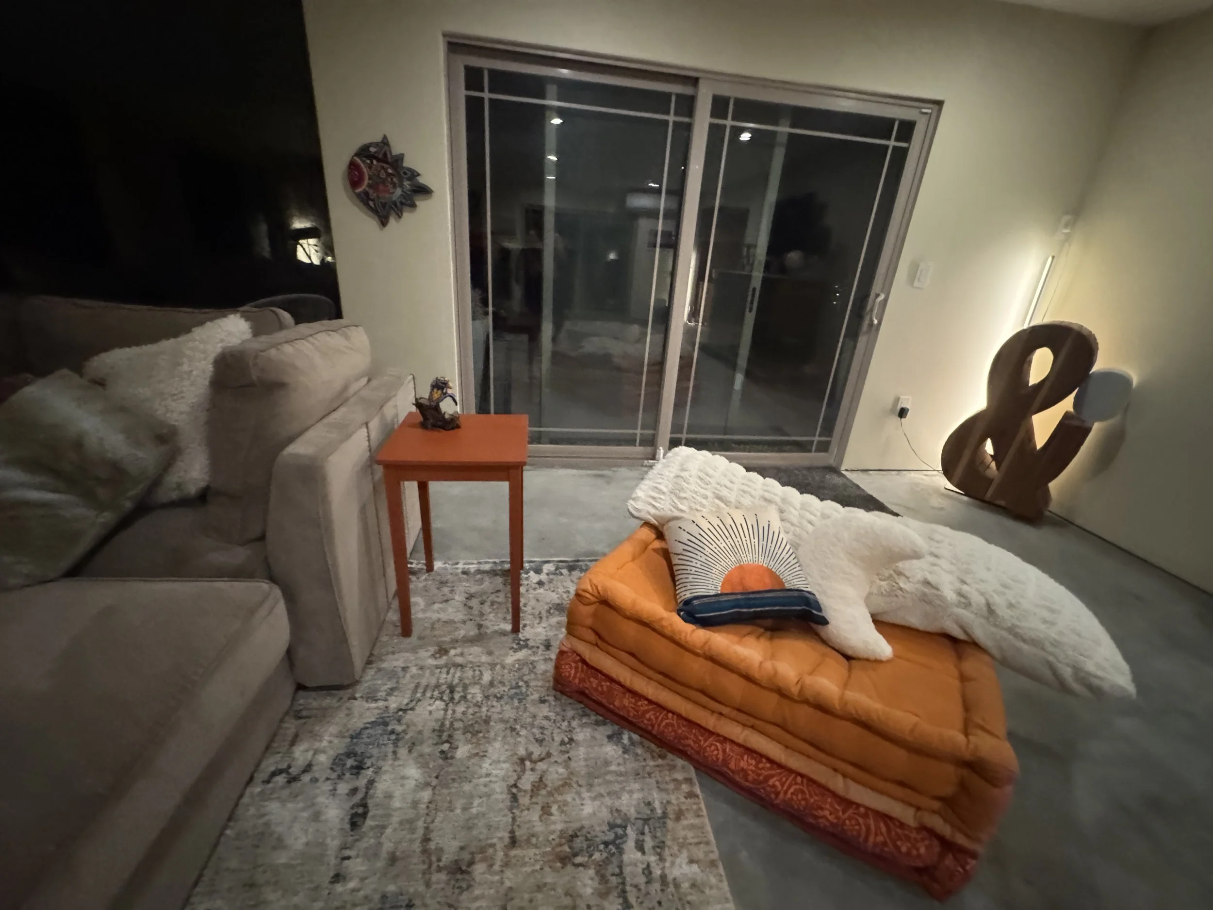 Living room with a beige couch on the left, a small orange table next to it, and a large sliding glass door leading outside. An orange tufted ottoman with pillows and throws on top sits in the center. A modern abstract wooden sculpture stands against