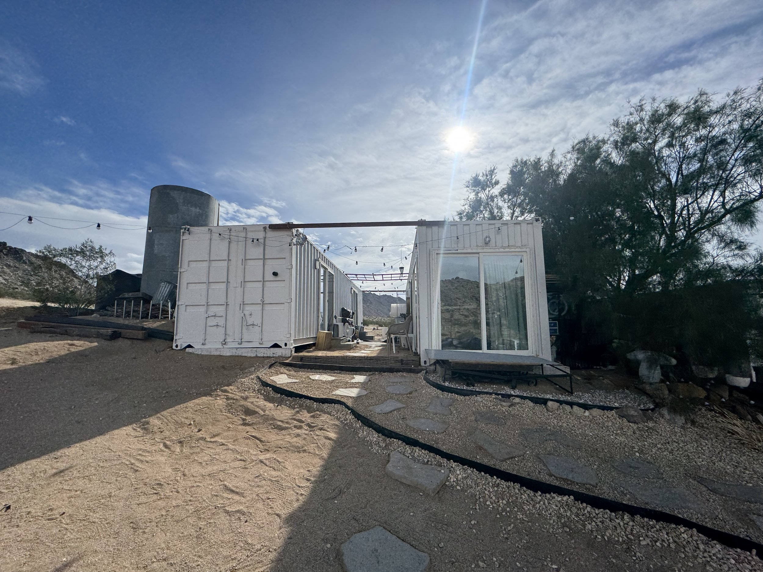 A makeshift outdoor living space created from shipping containers, with string lights overhead and desert landscaping, under a bright cloudy sky.