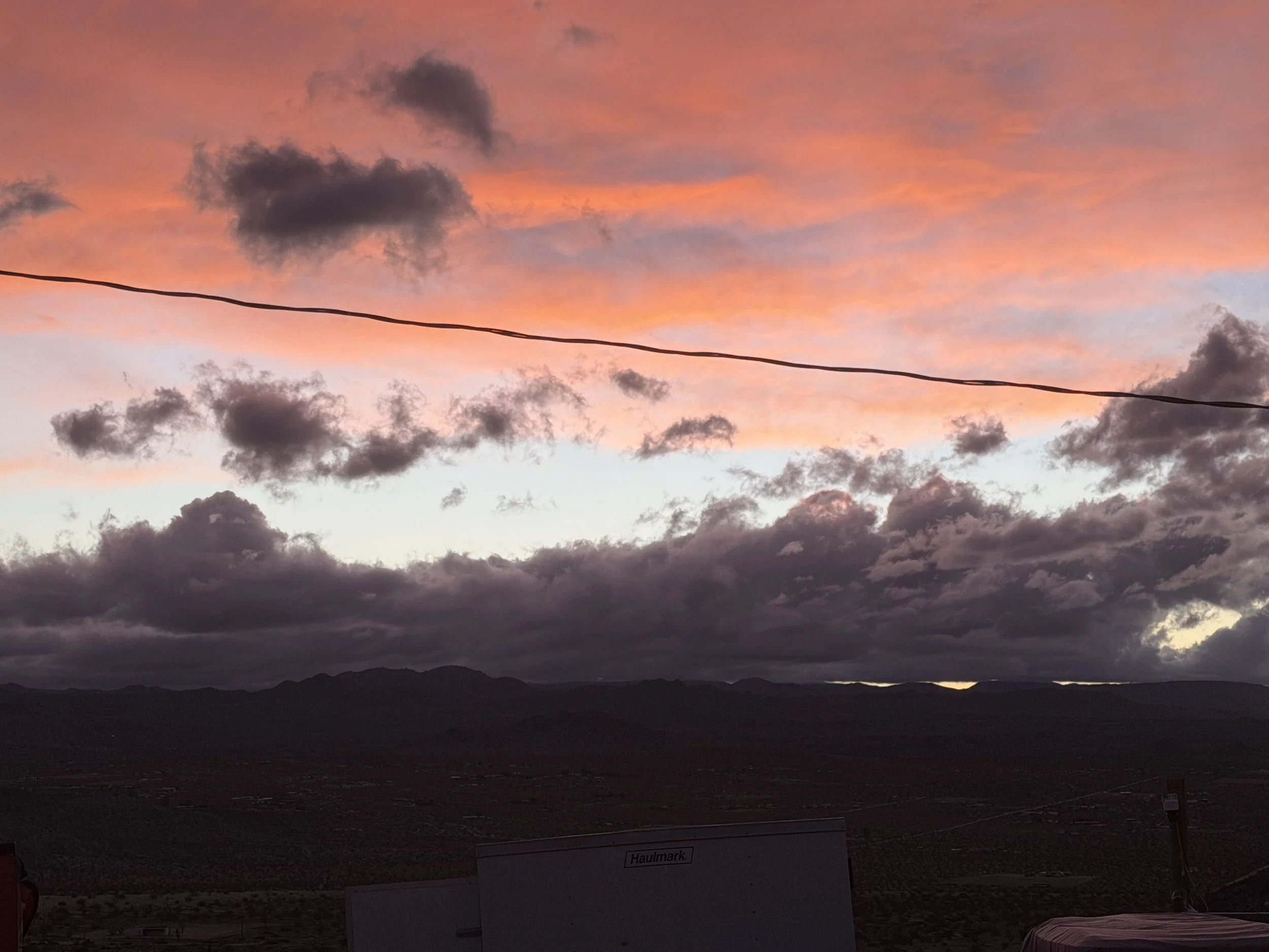 Sunset sky with pink, orange, and purple clouds over a dark mountainous landscape, with a power line running across the scene.