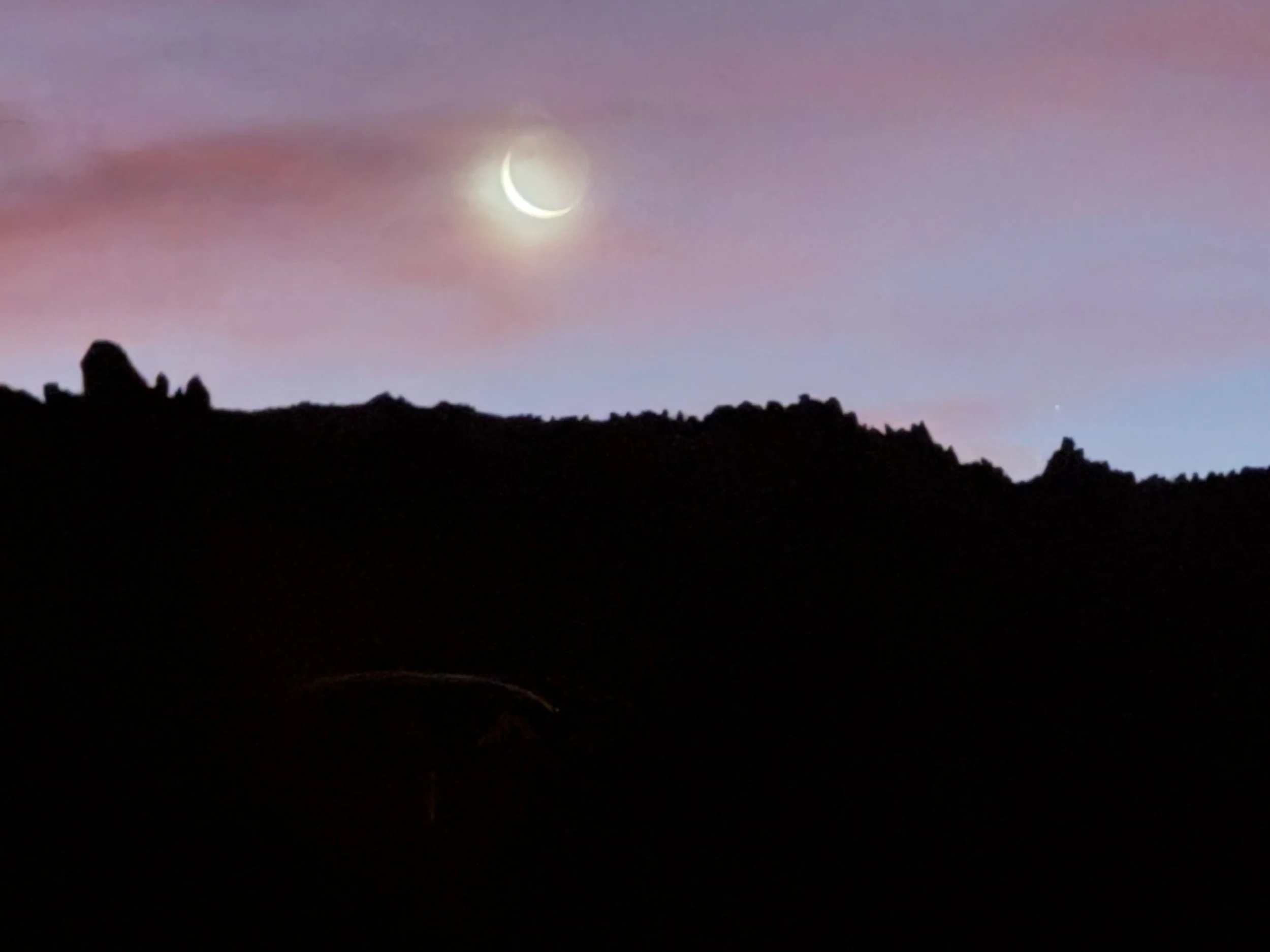 Nighttime landscape with a crescent moon in the sky, pink and purple clouds, and a dark silhouette of a hilly terrain with jagged peaks.
