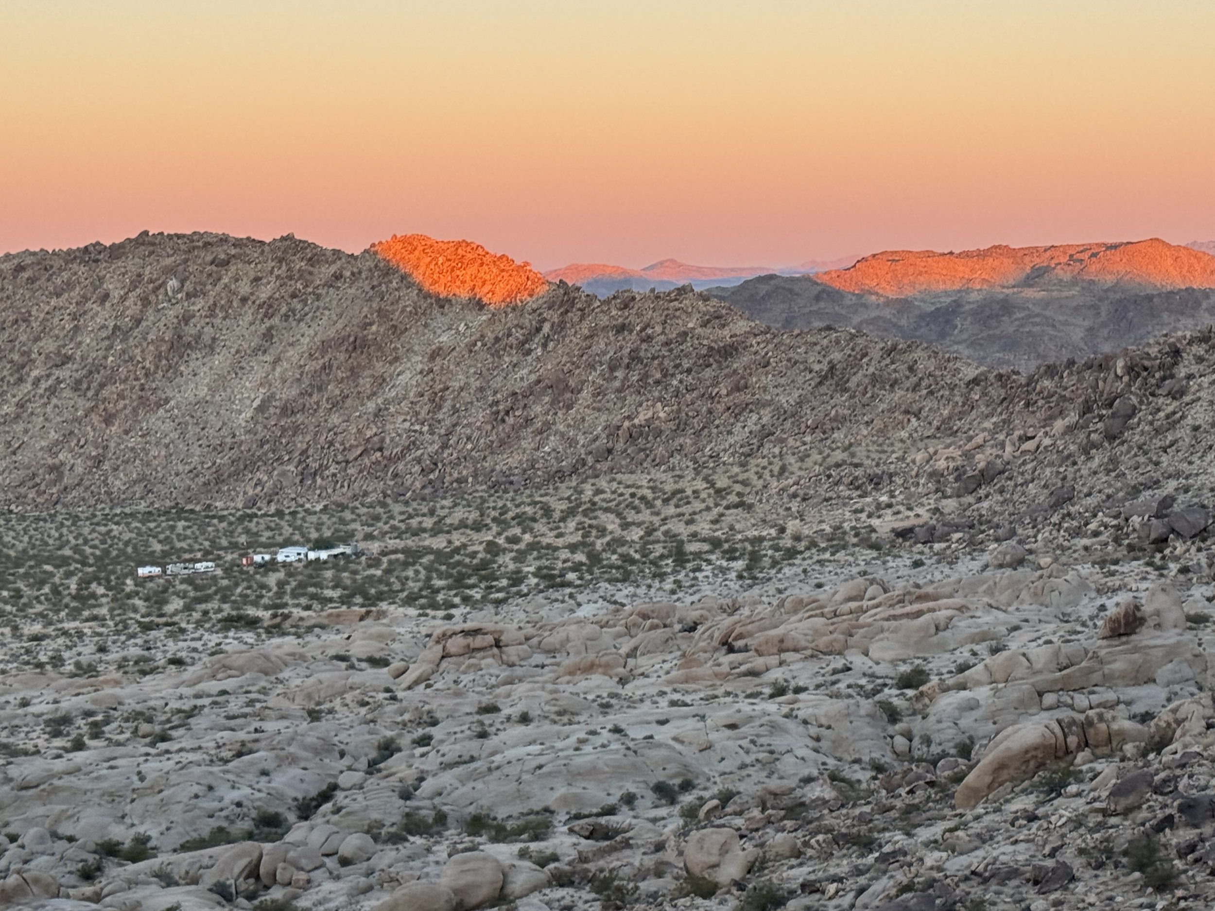 Desert landscape with rocky terrain and sparse vegetation, mountains in the background, and a cluster of small buildings in the valley at sunset.