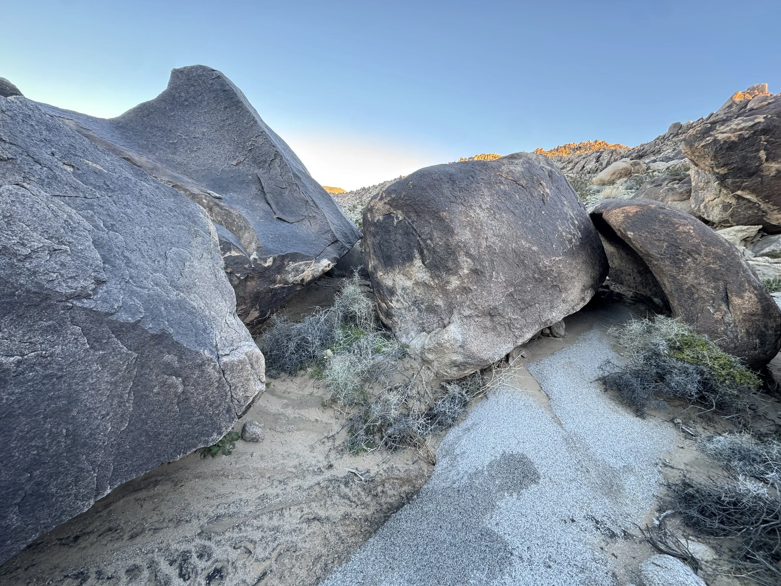 Large boulders on a desert landscape with sandy ground and sparse dry bushes under a clear blue sky.