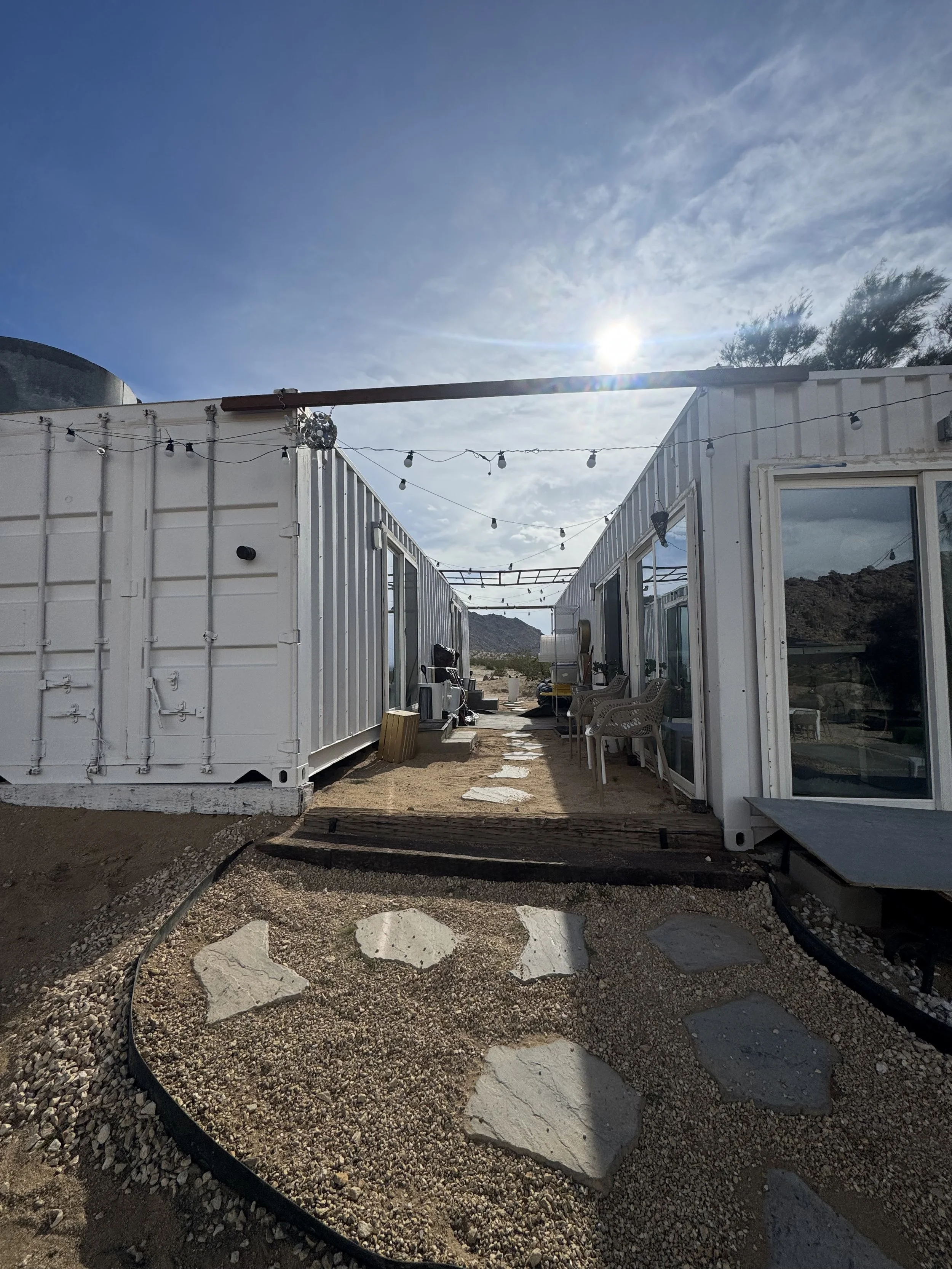 Two white shipping container buildings with string lights, outdoor furniture, and a stone pathway on desert terrain, with mountains and a blue sky in the background.