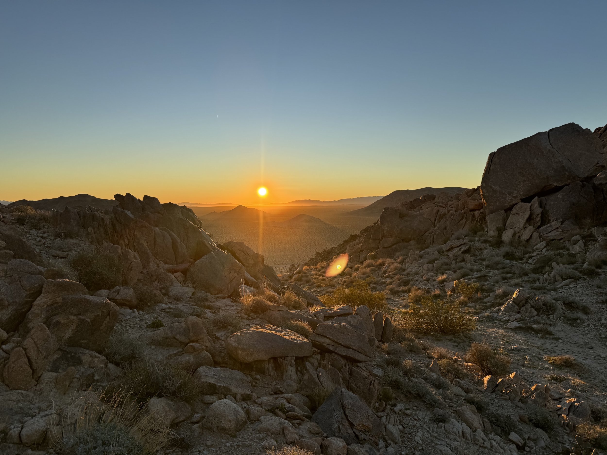 Sunset over a rocky desert landscape with scattered bushes and distant mountains.