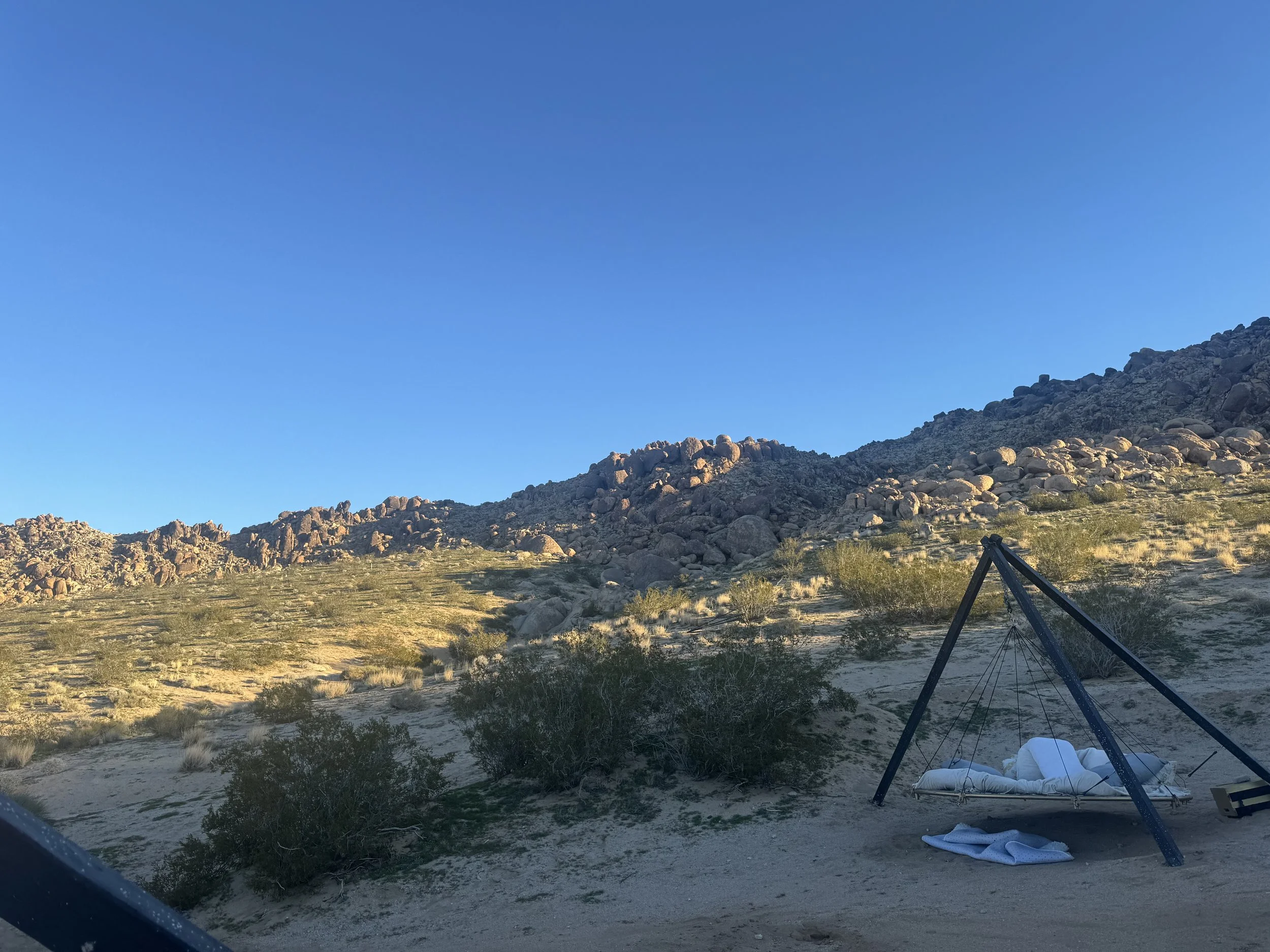 A desert landscape with rocky hills in the background under a clear blue sky. In the foreground, there is a black A-frame swing set with a white cushion or bed hanging from it, with some white towels or cloth underneath.