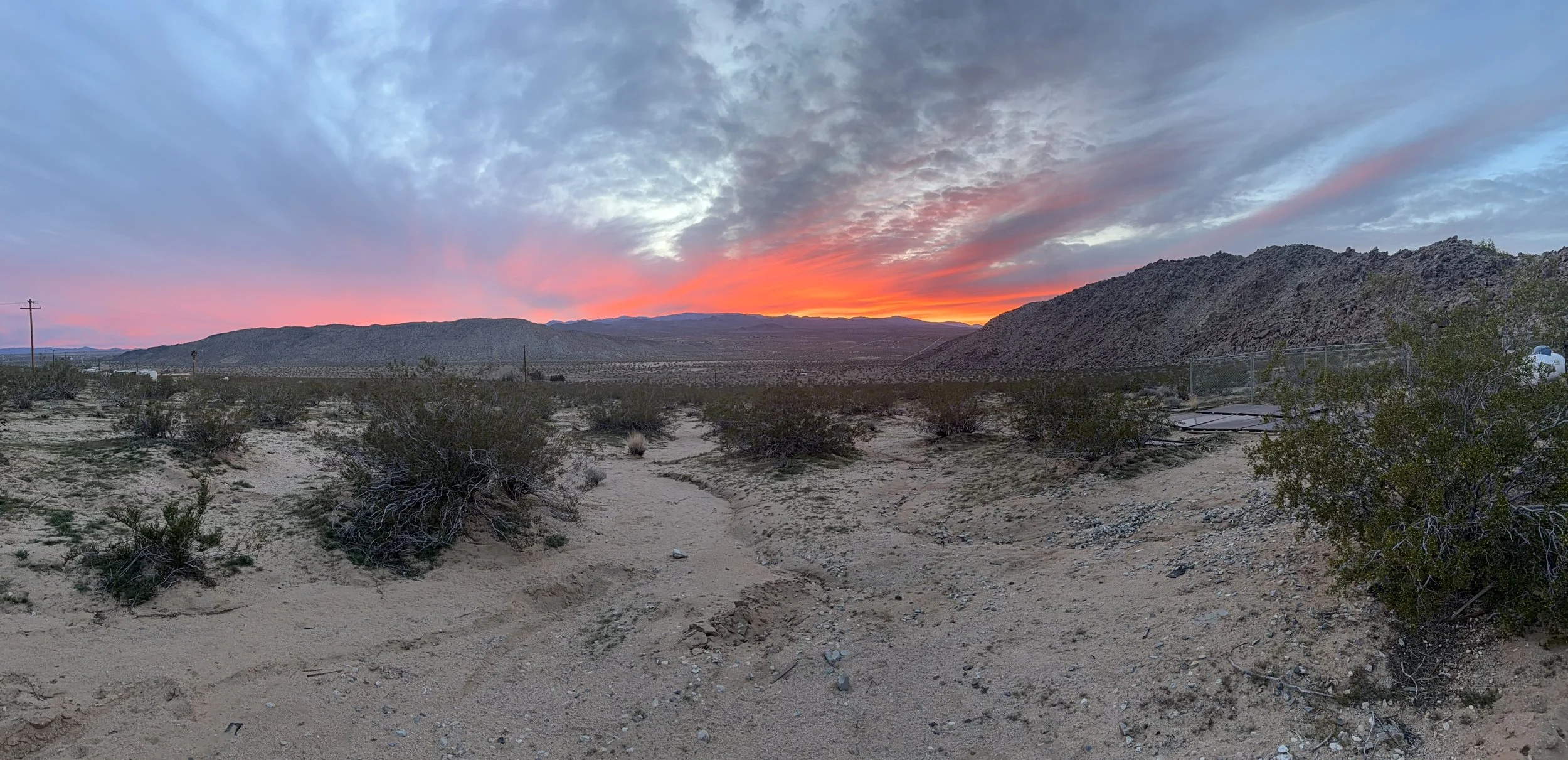 Desert landscape at sunset with pink, orange, and gray clouds over distant mountains, sparse desert vegetation, dirt ground, and a fence on the right side.
