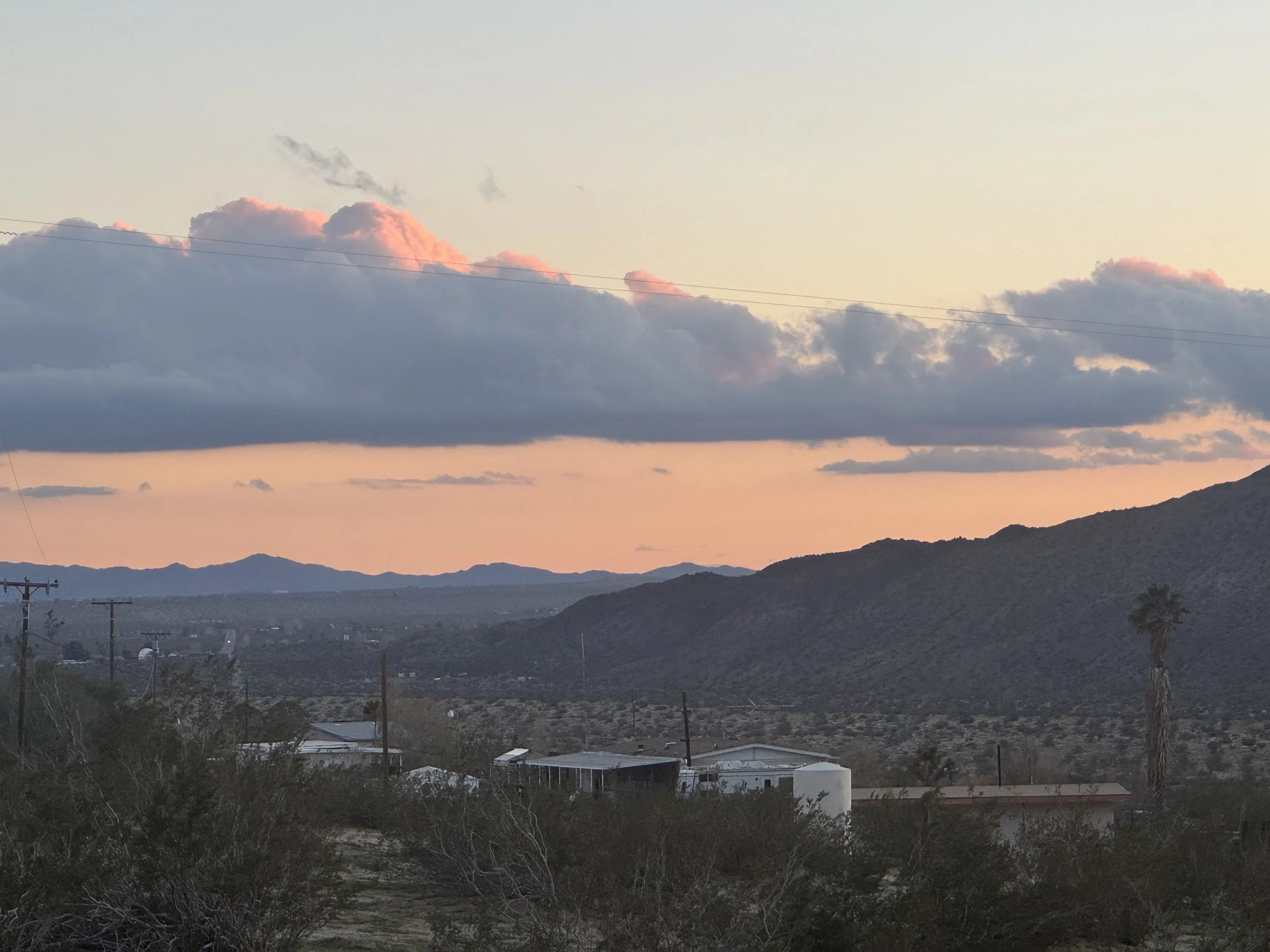 Scenic view of desert landscape with mountains, a few buildings, trees, and a large cloud formation during sunset.