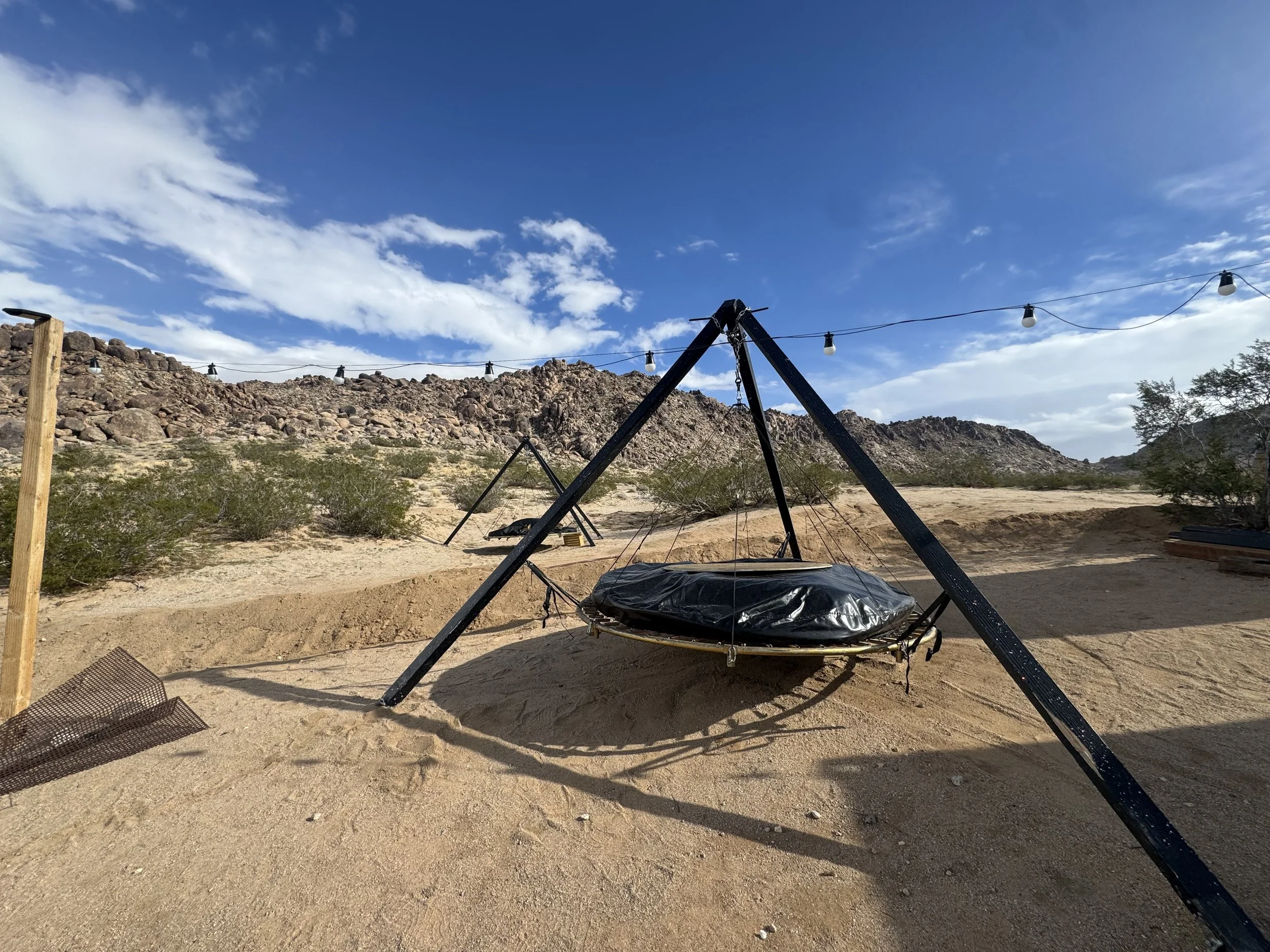 Outdoor swing with black cover suspended from black tripod structure in a desert landscape with bushes, rocky hills, and string lights overhead during daytime.