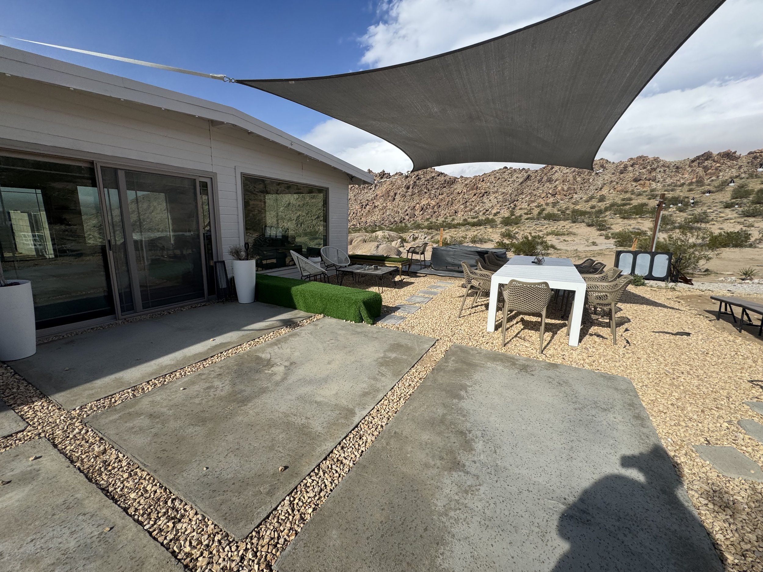 Desert outdoor patio with grilled sitting area, white table and chairs, shaded by a large fabric sunshade, with rocky hills in the background.