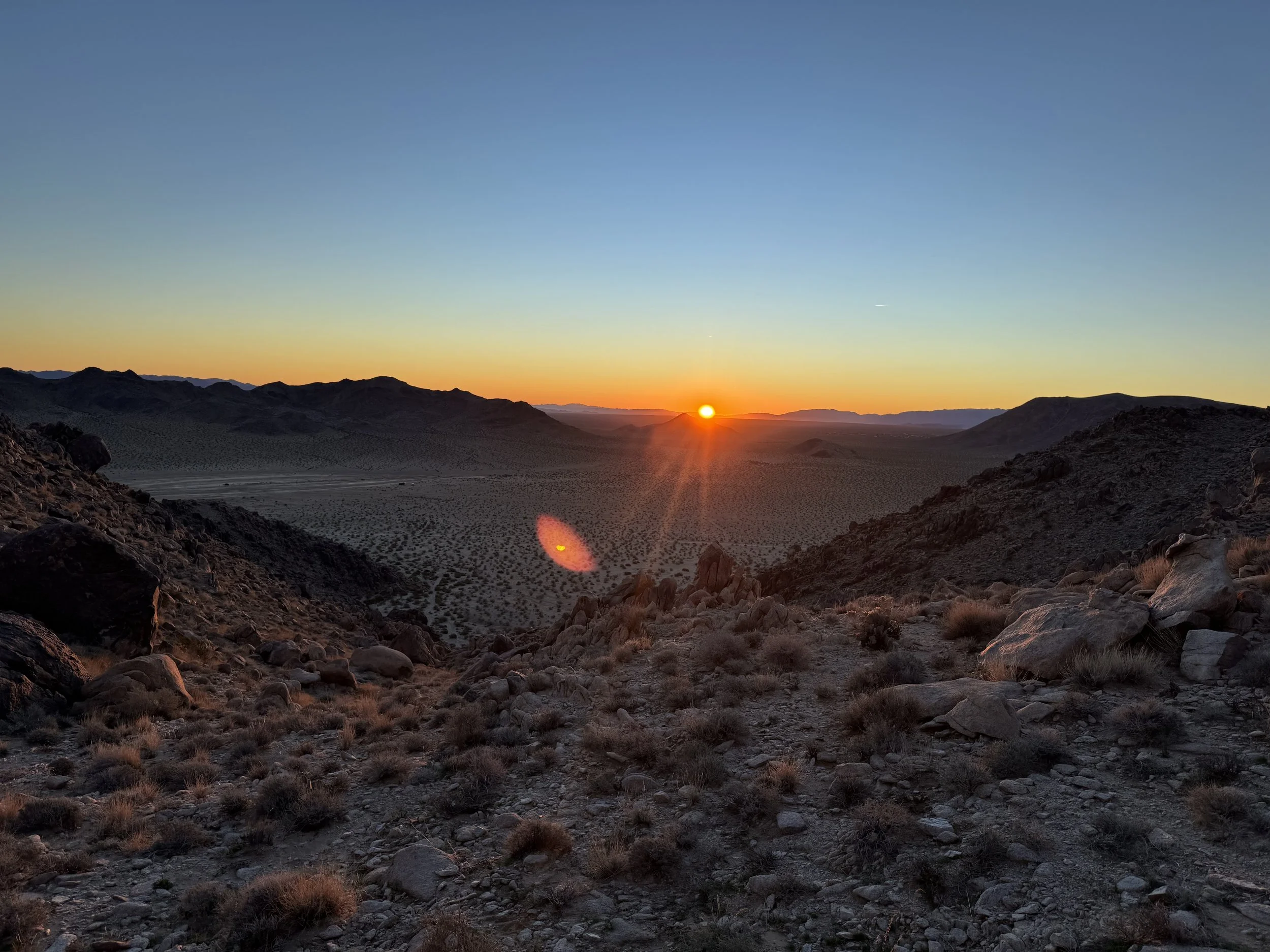 Desert landscape at sunset with rocky terrain and distant mountain ranges.